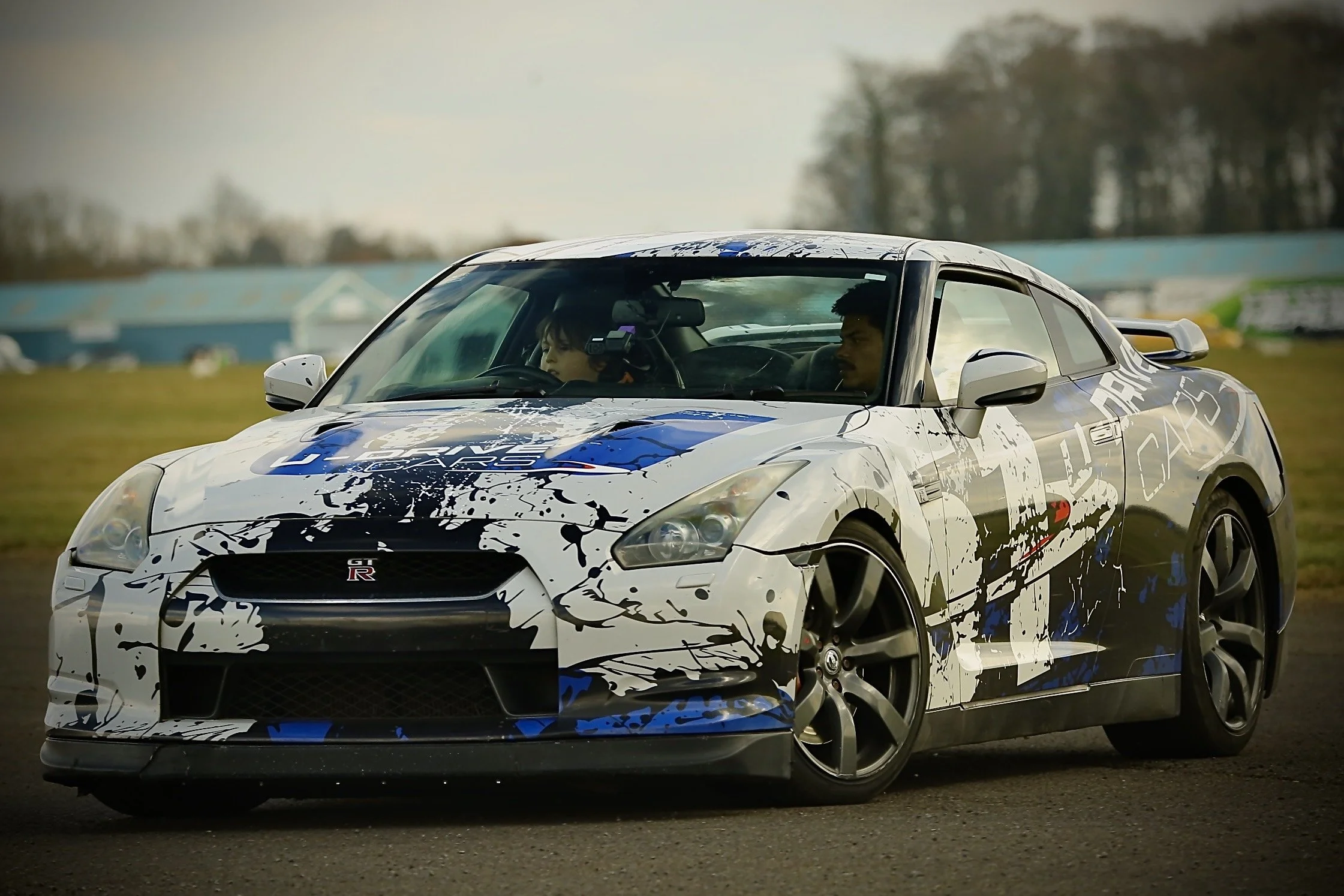A white sports car with blue and black splatter paint design, the Nissan GT-R logo visible on the front, on a race track with two people inside, a man and a young boy, both wearing seat belts, with the boy in the passenger seat.