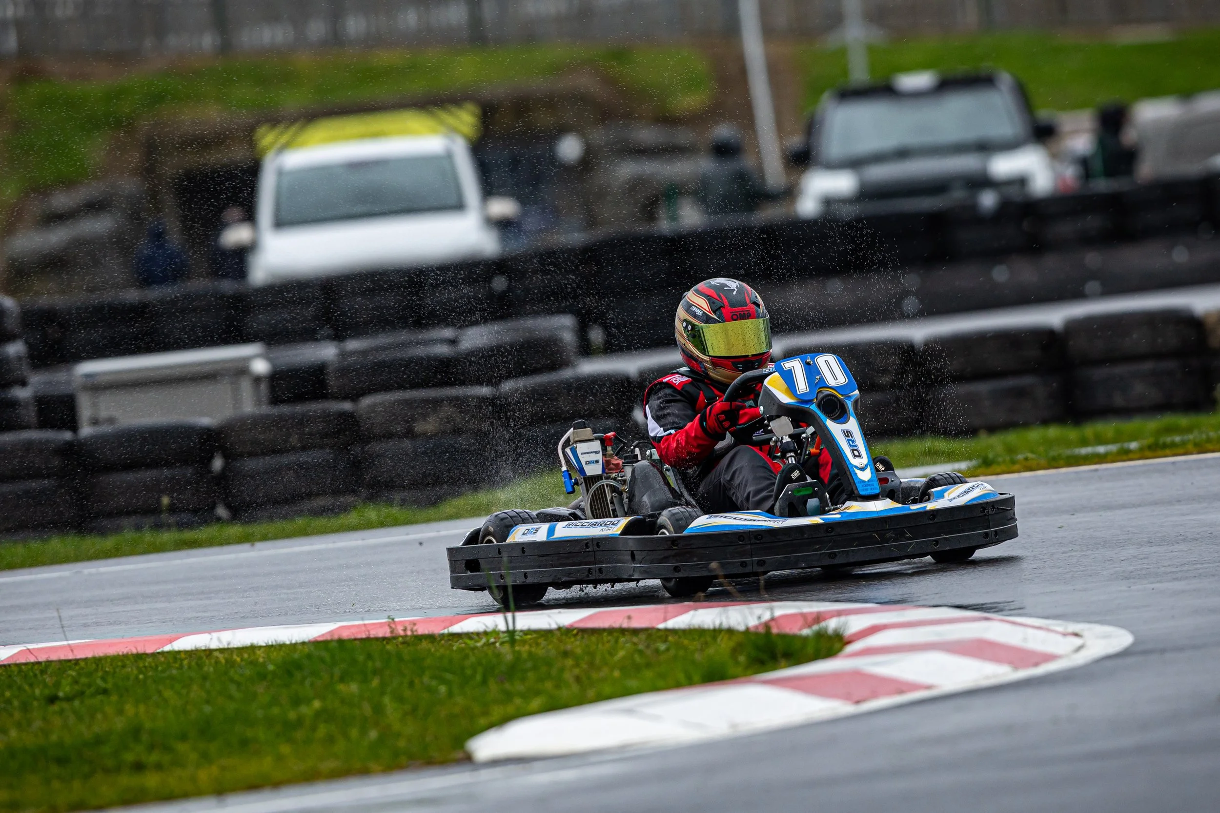 A go-kart driver in red and black gear and a helmet racing on a track, with blurred cars and a guardrail in the background, rain droplets on the camera lens.