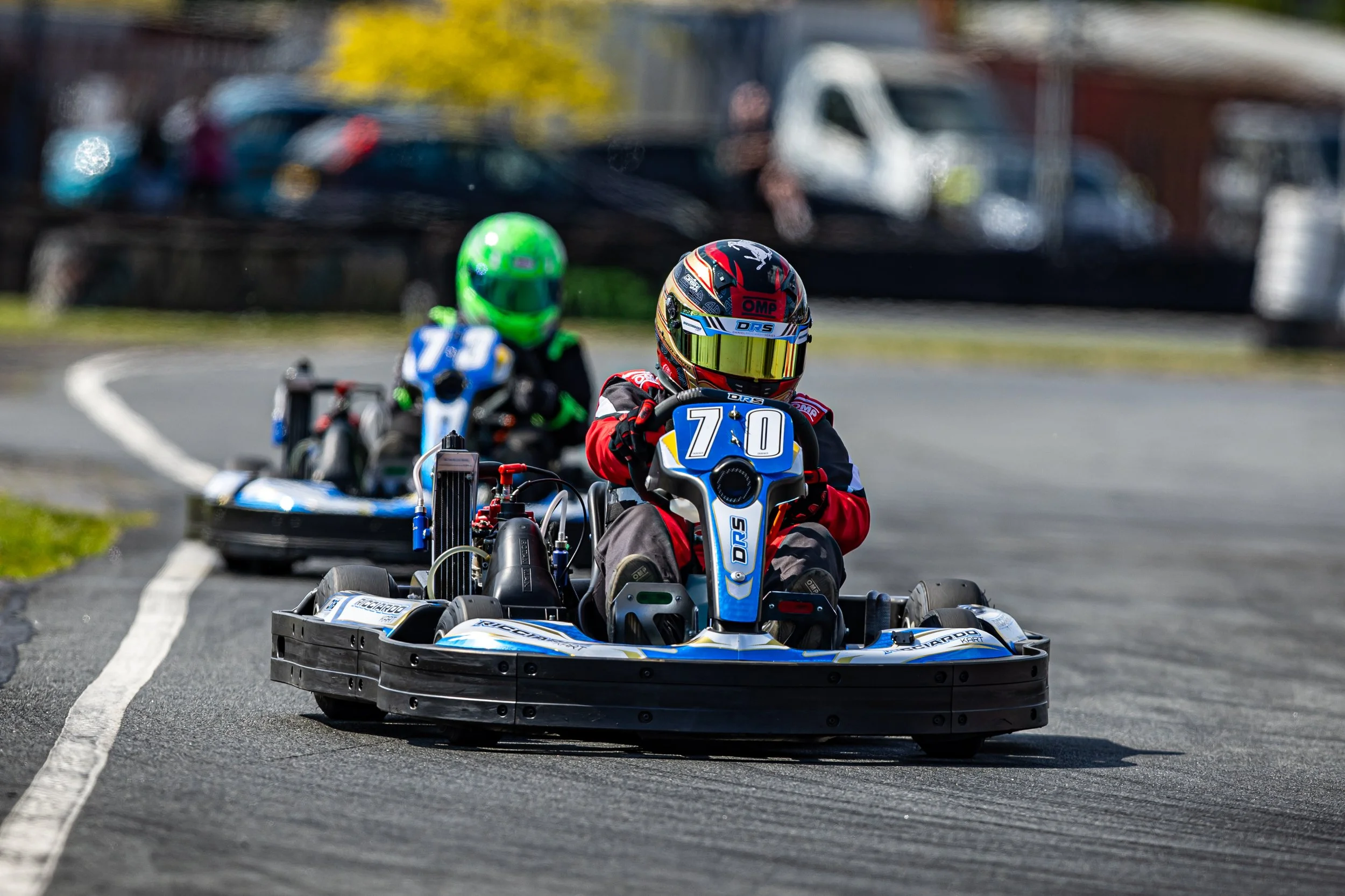 Two go-kart racers wearing helmets and racing suits on a track during a race.