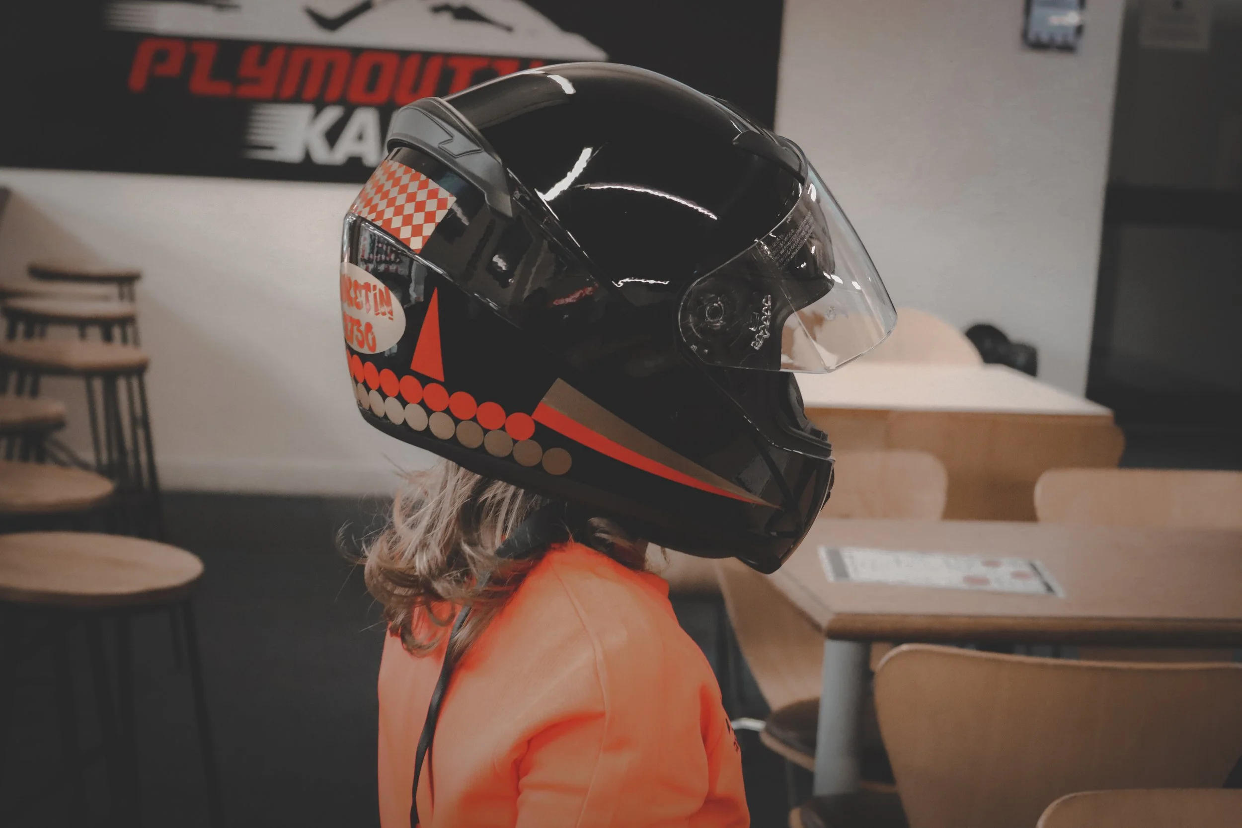 Child wearing a black motorcycle helmet with orange and white designs, sitting indoors.