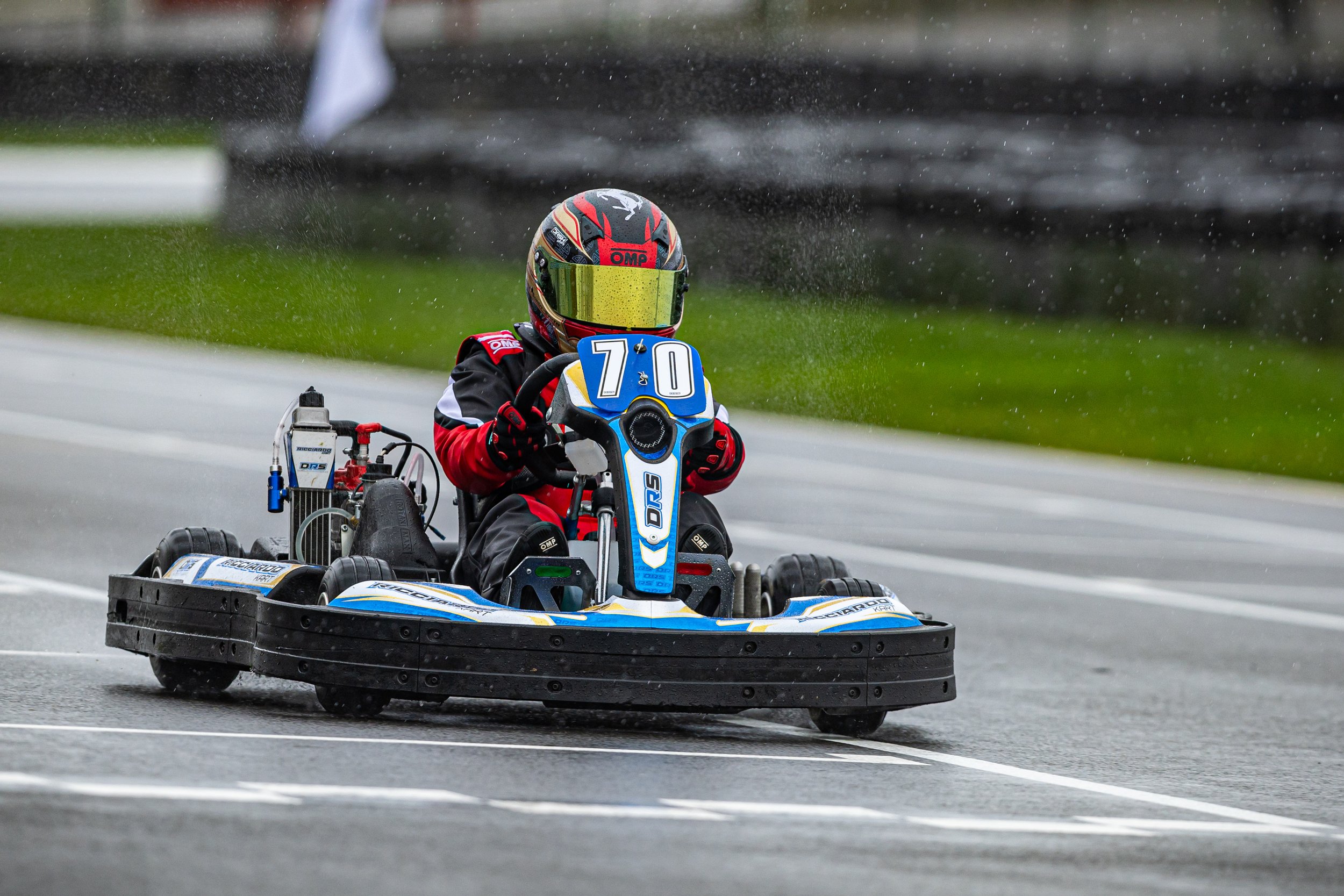 A person racing a go-kart on a paved track, wearing a black helmet with red and yellow accents and a reflective visor, a black and red racing suit, and gloves, with a blurred background of a grassy area and track barriers.