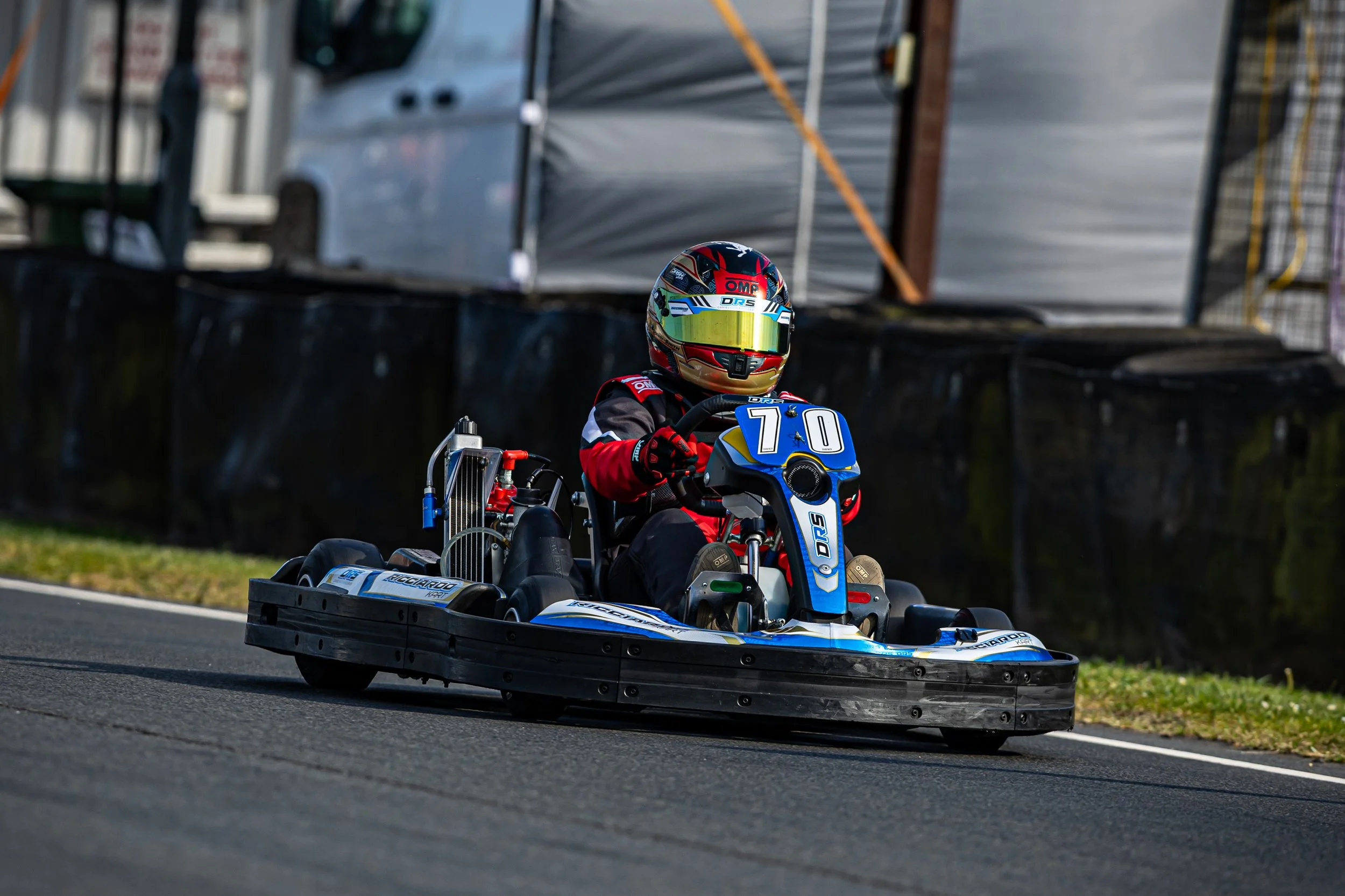 A go-kart racer in a red, black, and blue racing suit and a colorful helmet, sitting in a blue and white go-kart on a race track.
