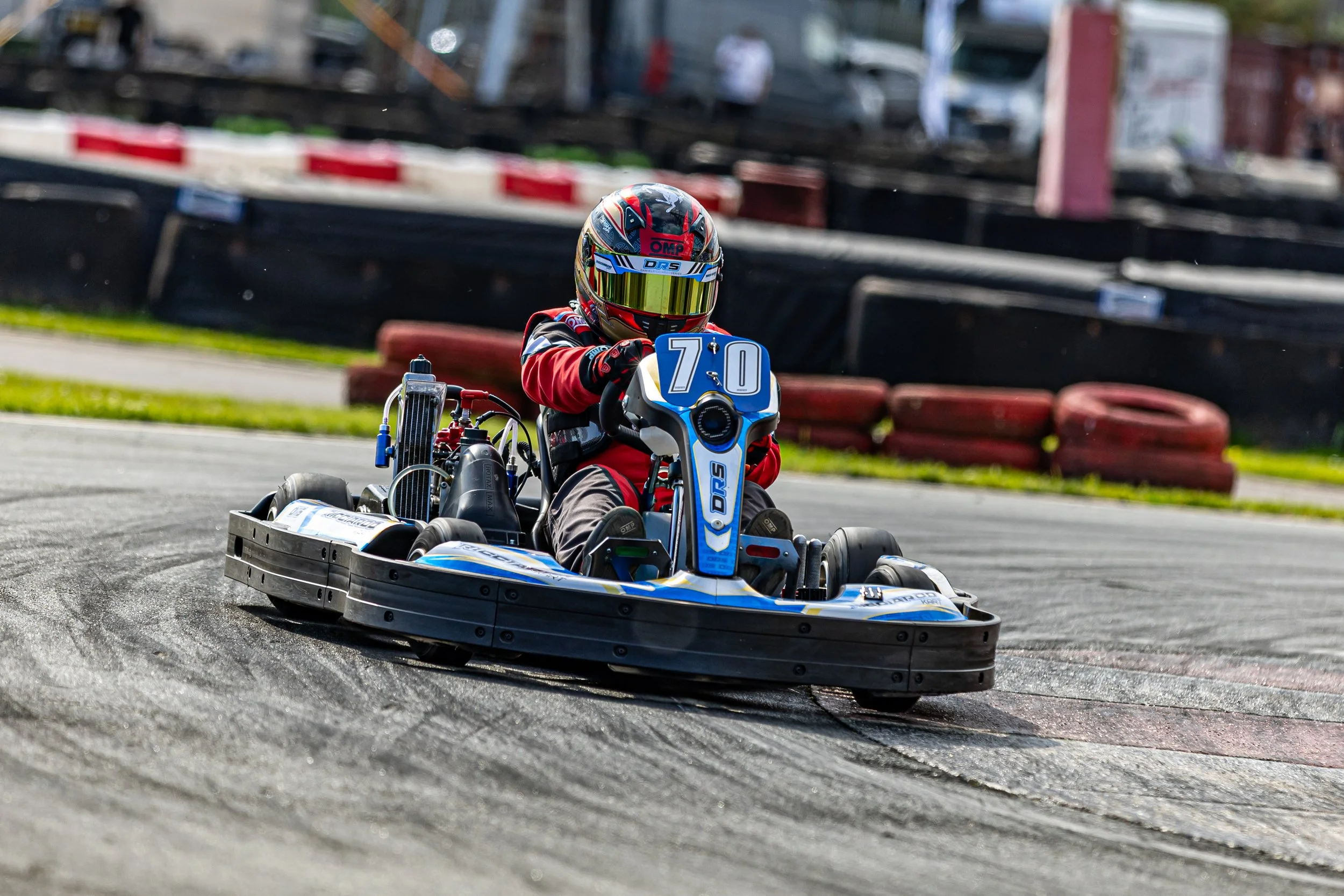 A person driving a go-kart on a race track, wearing a red and black racing suit and a helmet with a reflective visor.