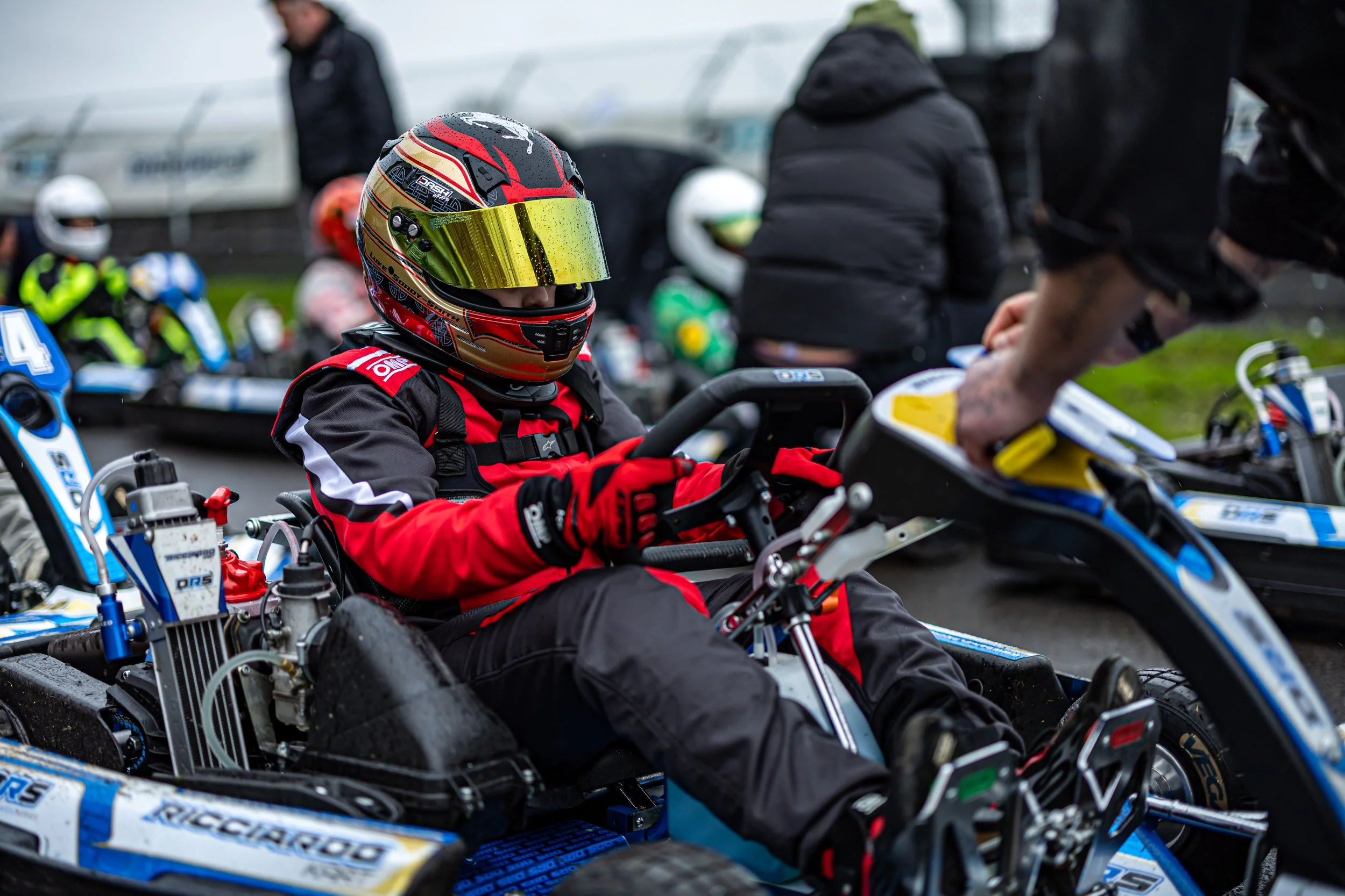 Close-up of a race car driver in a red and black racing suit and a colorful helmet sitting in a go-kart, with other racers and team members in the background.