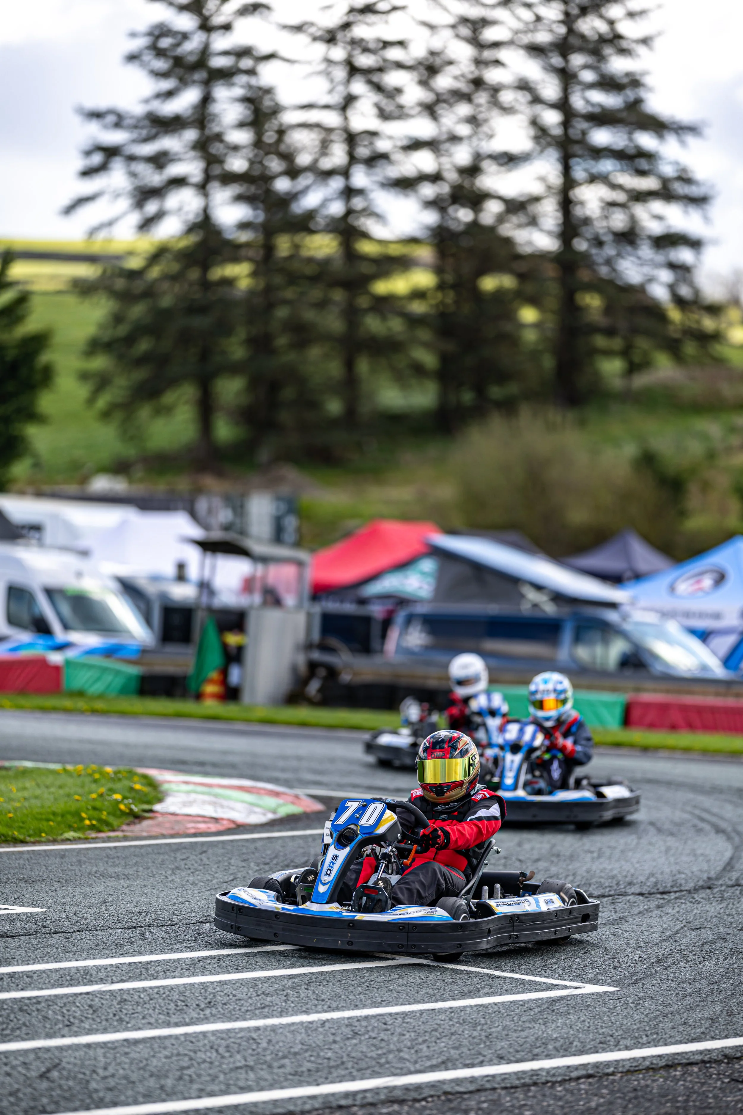 Four drivers in go-karts racing on an outdoor track during daytime, with colorful tents and trees in the background.