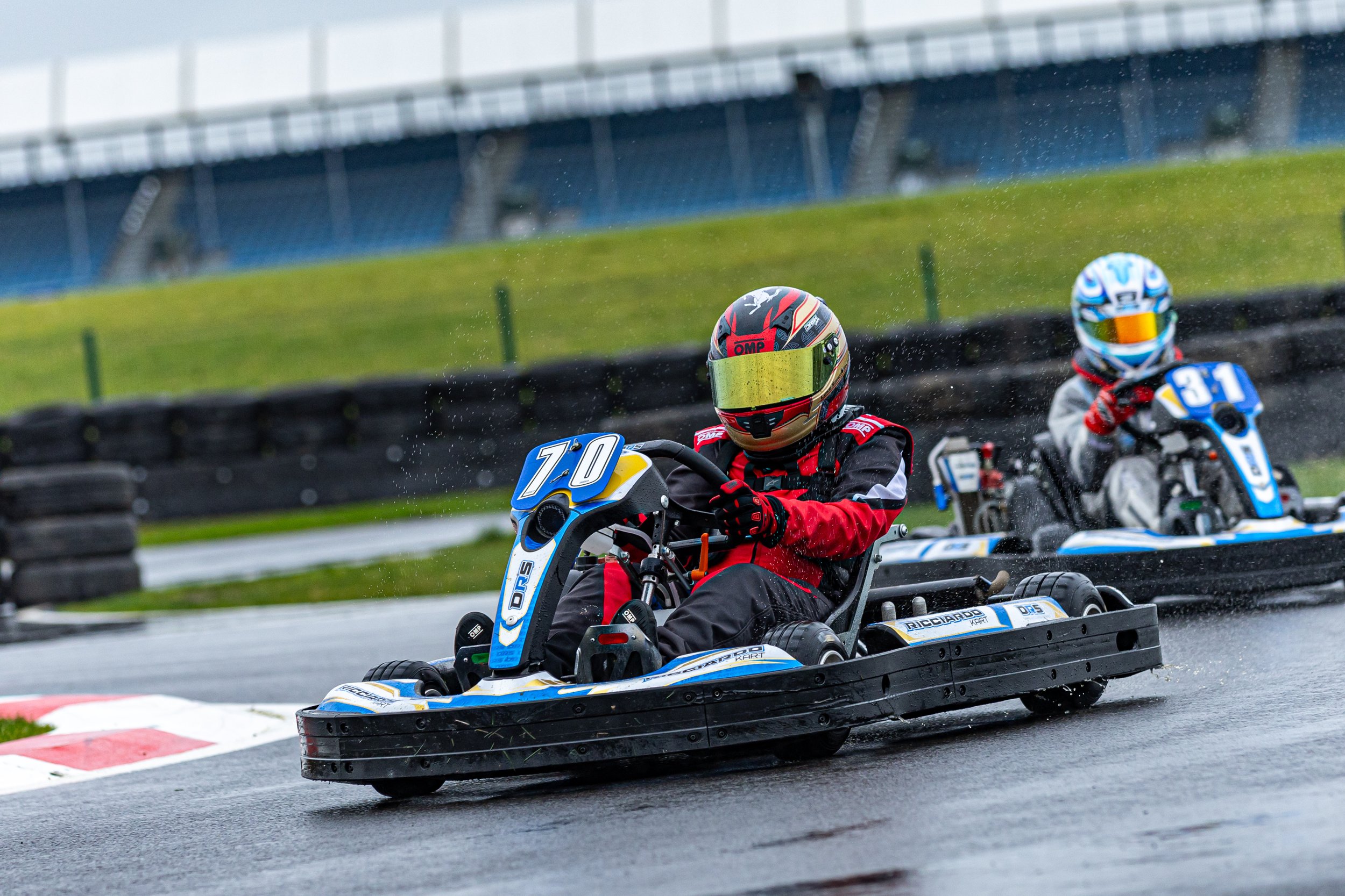 Two go-kart racers wearing helmets and racing suits on a wet track, with one kart marked with the number 70 and the other with the number 81.