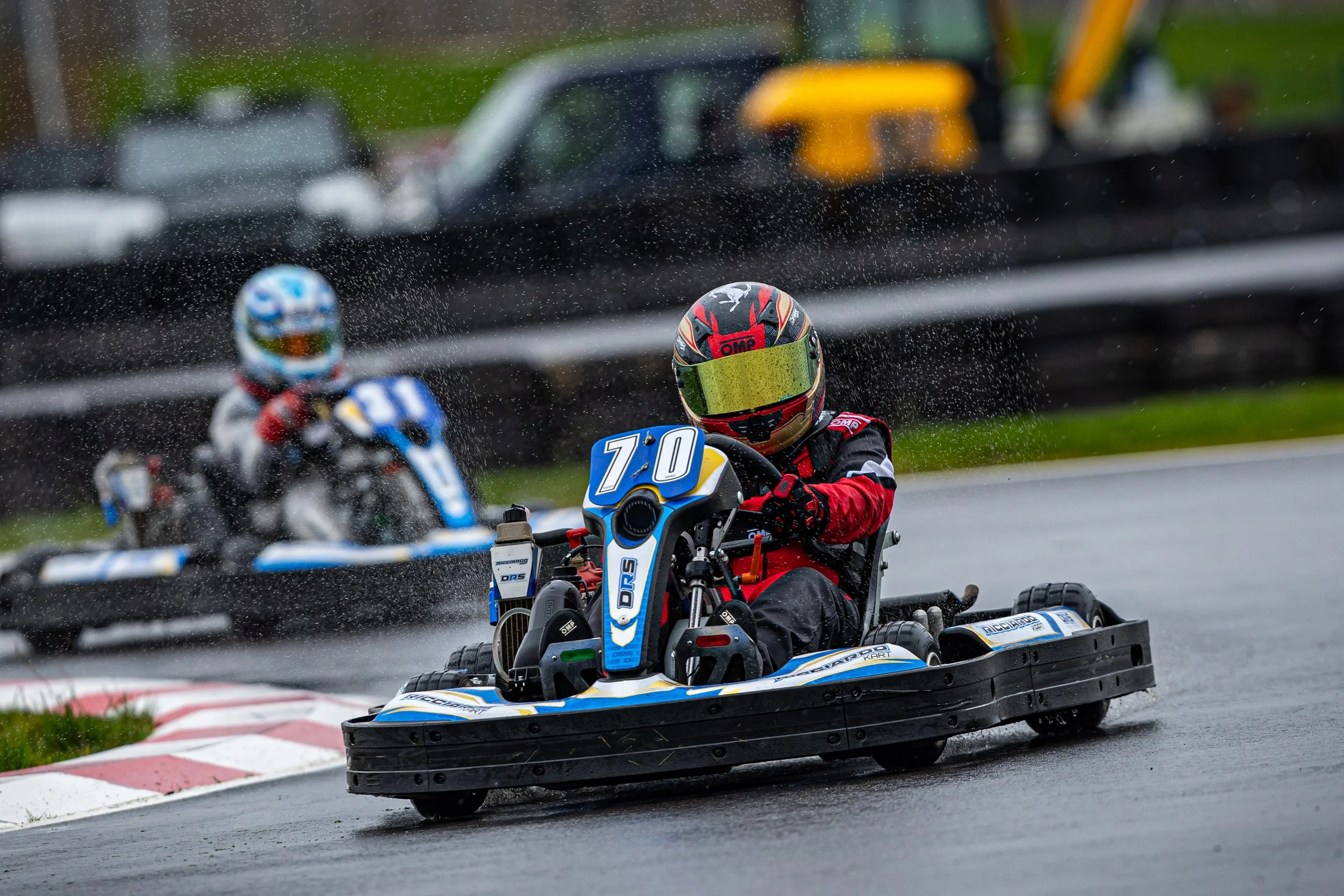 Two go-kart racers wearing helmets and racing suits on a wet track, with rain droplets splashing and blurred vehicles in the background.