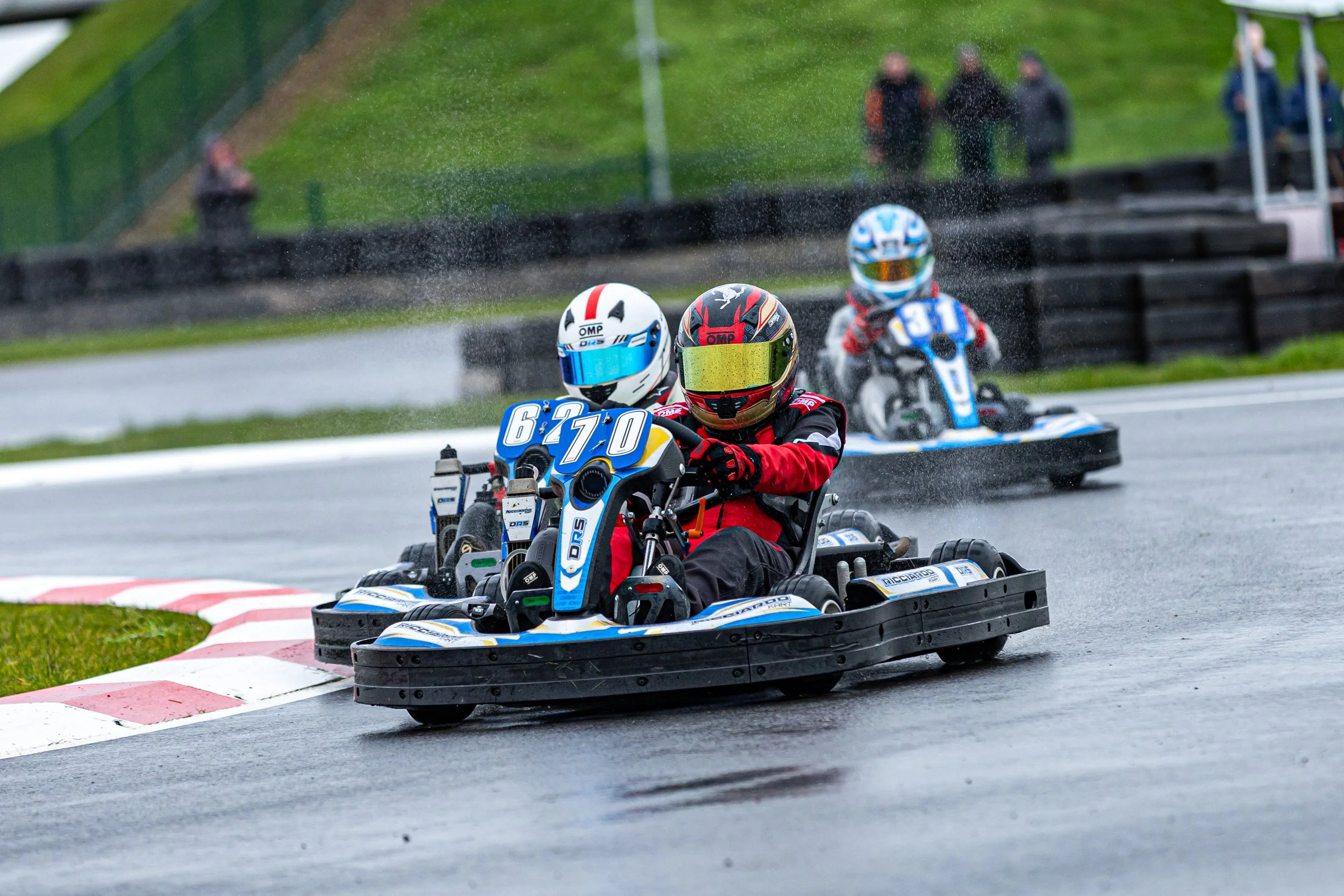Three go-kart racers wearing helmets and racing suits on a track during rainy weather, navigating a turn.