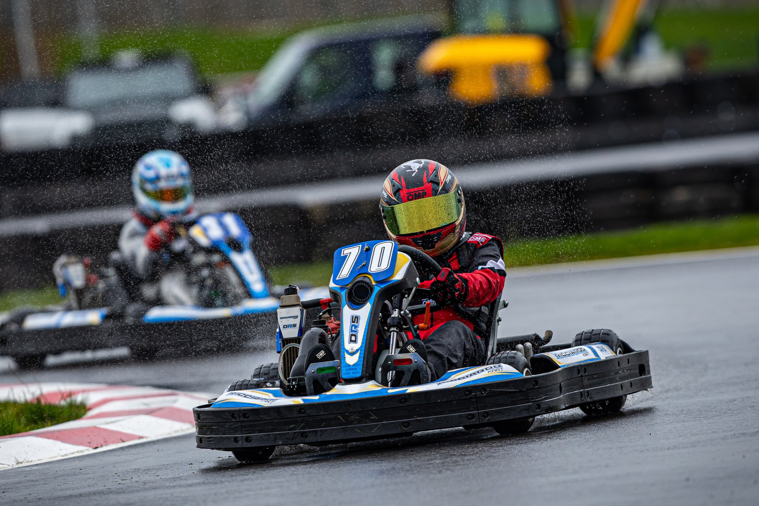 Two go-kart racers navigating a wet track with water spray visible, one in focus wearing a black and red helmet and red jacket, the other blurred in the background.