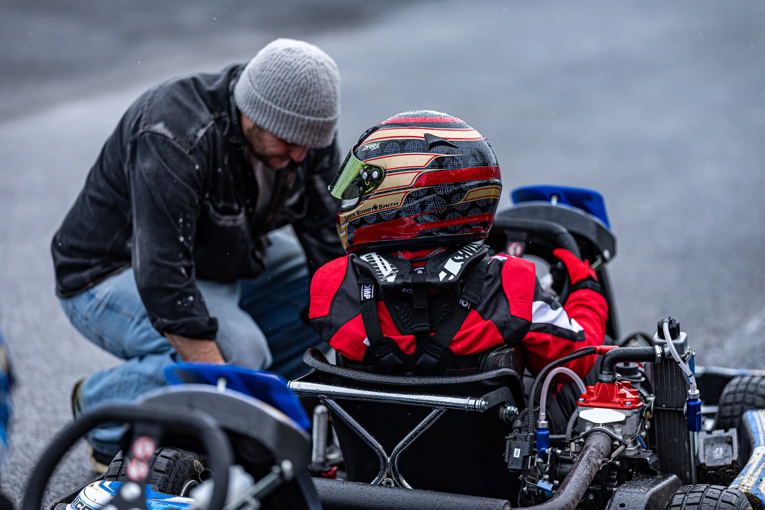 A man in a gray beanie and black jacket helps a person in race gear and helmet who is sitting on a racing wheelchair, likely preparing for a race or a race-related activity.