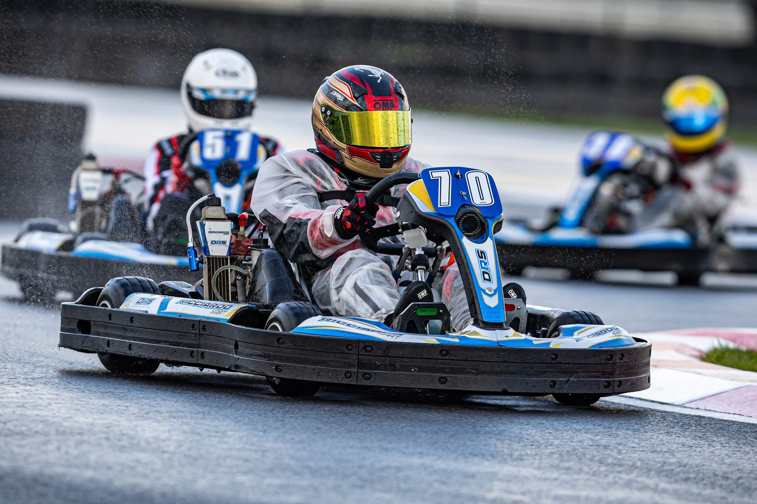 A go-kart race car speeds along a wet track, with three racers wearing helmets and racing suits visible, highlighting the excitement of the race.