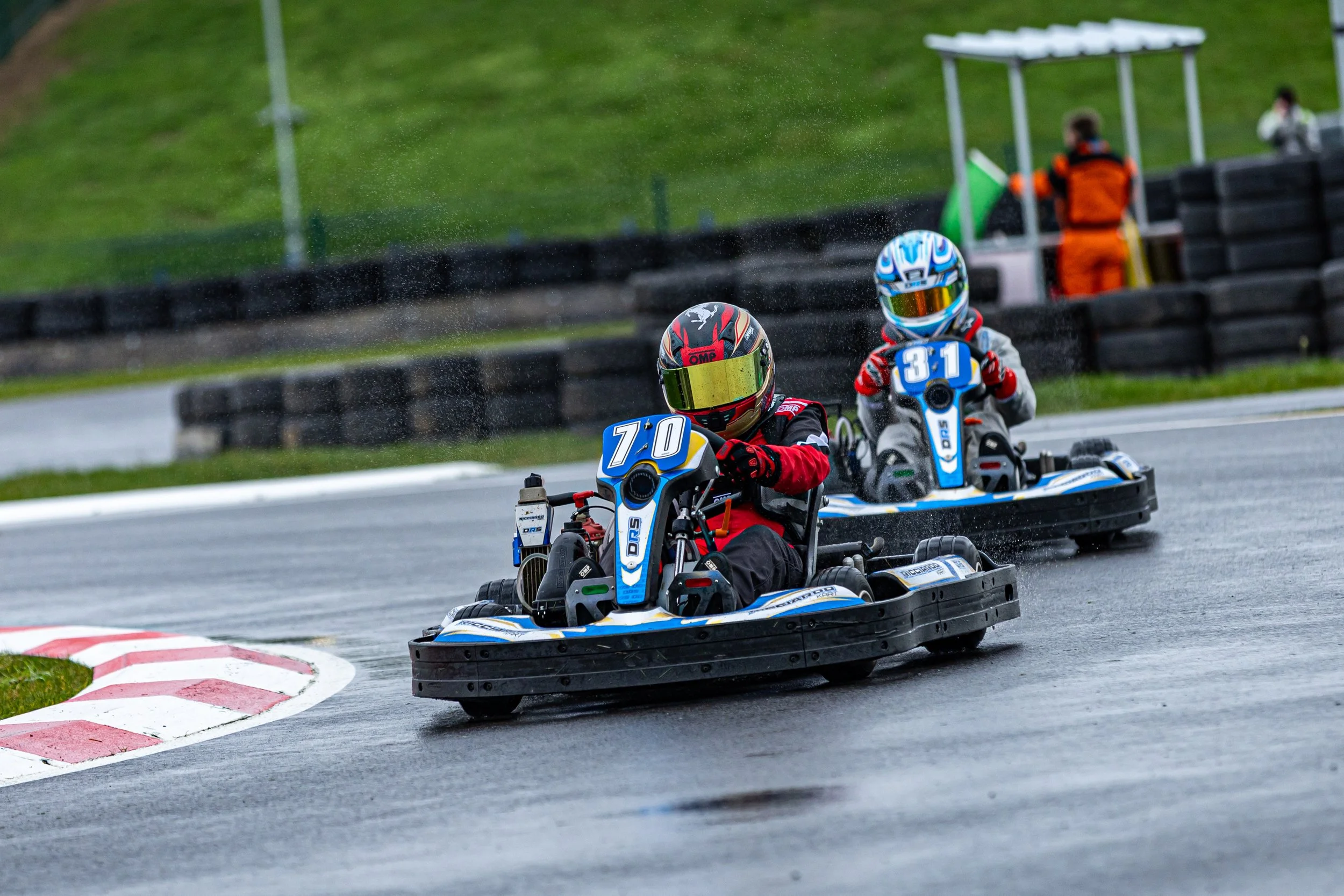 Two go-karts racing on a wet track with drivers wearing helmets and racing suits, and a person in orange and black in the background.