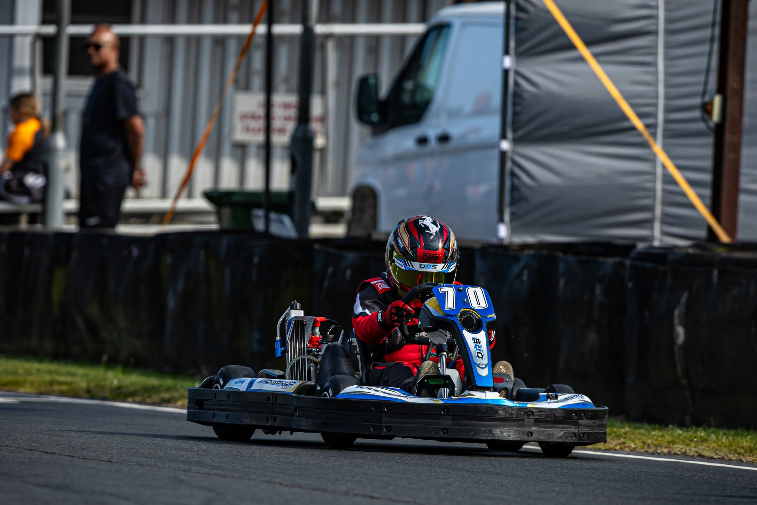 A go-kart driver wearing a helmet and racing suit driving on a race track, with a fade-out background showing a man and a boy standing near a truck and a fence.