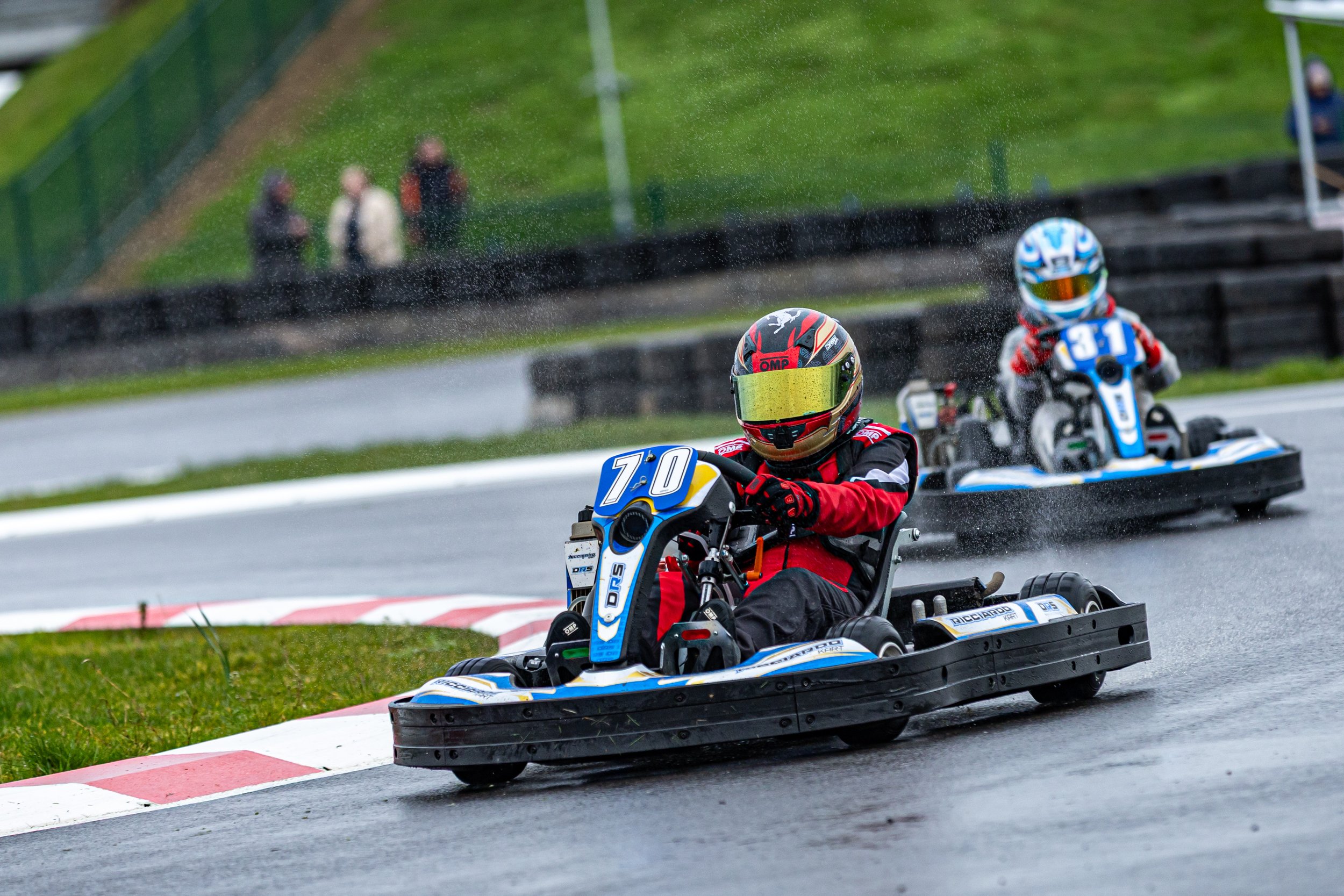 Two go-kart racers wearing helmets and racing suits speed around a track, with onlookers visible in the background.