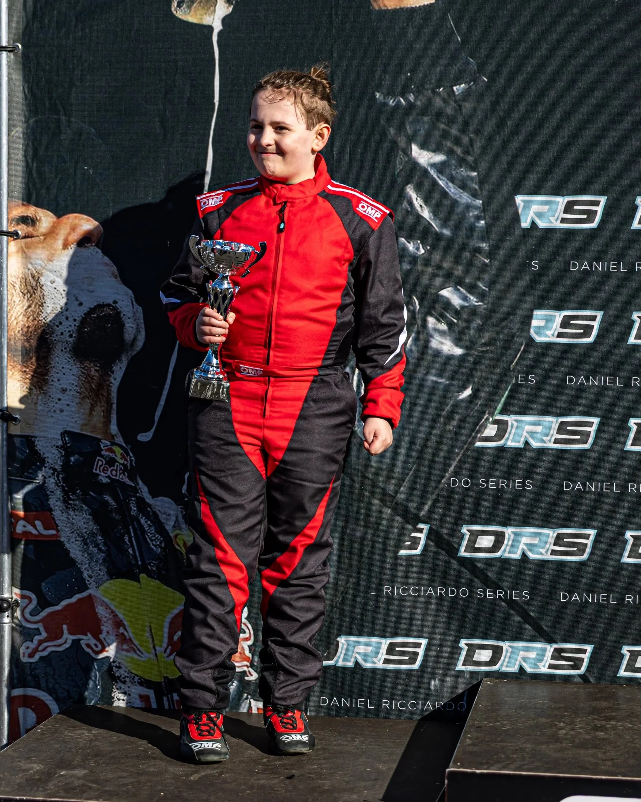 Young person in racing suit holding a trophy on a winner's podium at a racing event, with a black backdrop featuring logos and sponsor names.