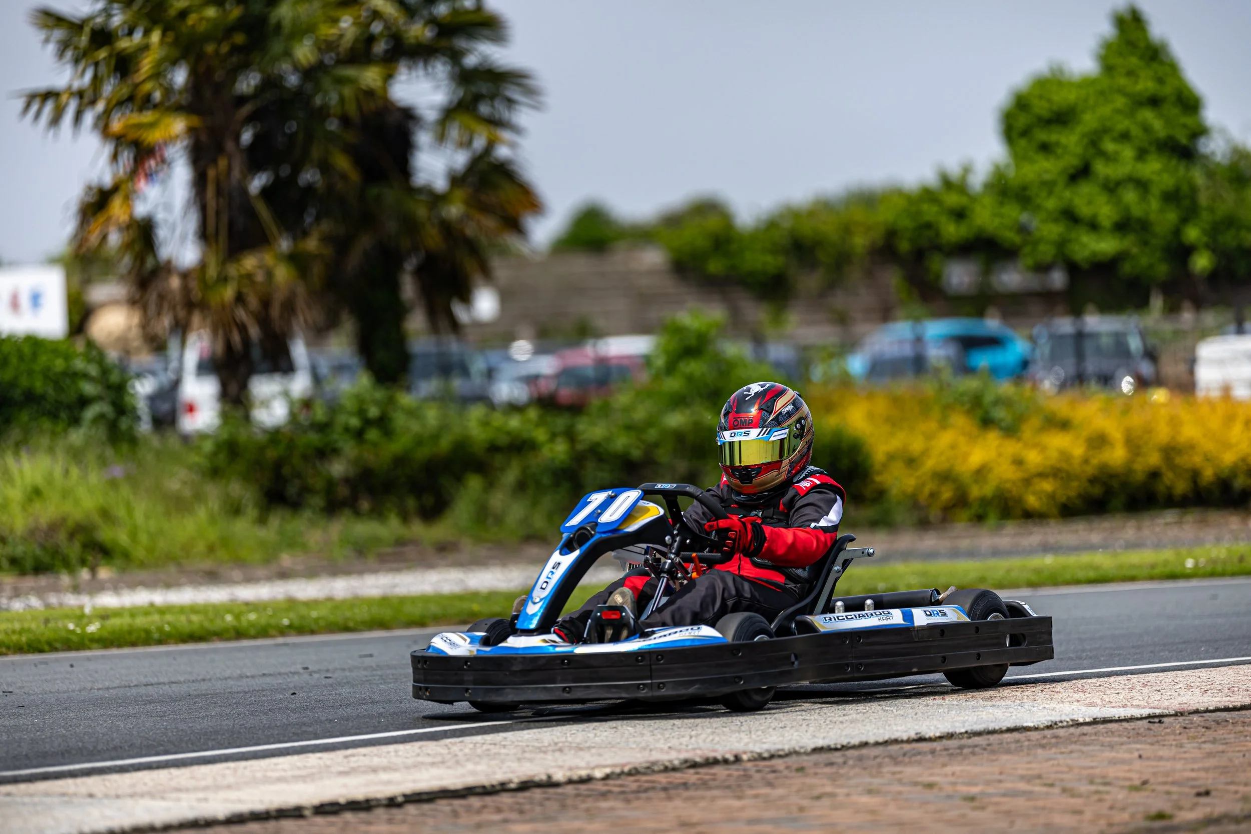 A person in a racing helmet and protective gear driving a go-kart on a paved track with trees and parked cars in the background.