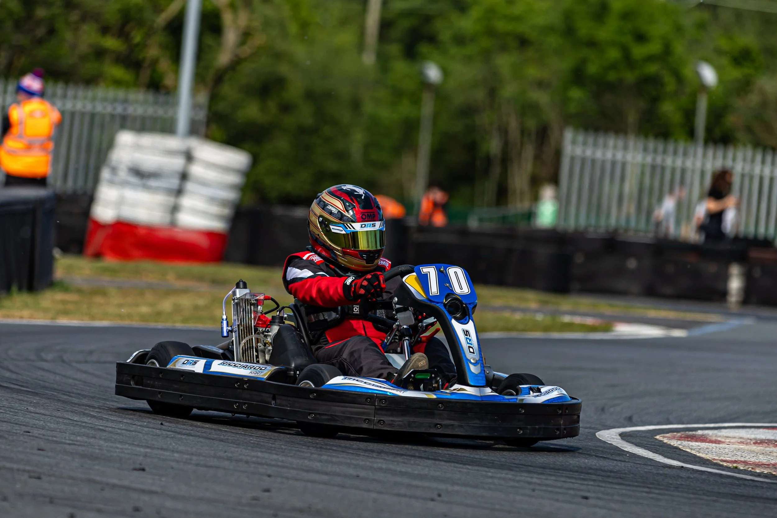 Go-kart racer wearing a red and black racing suit and a red, black, and gold helmet, driving on a race track with tires and a fence in the background.