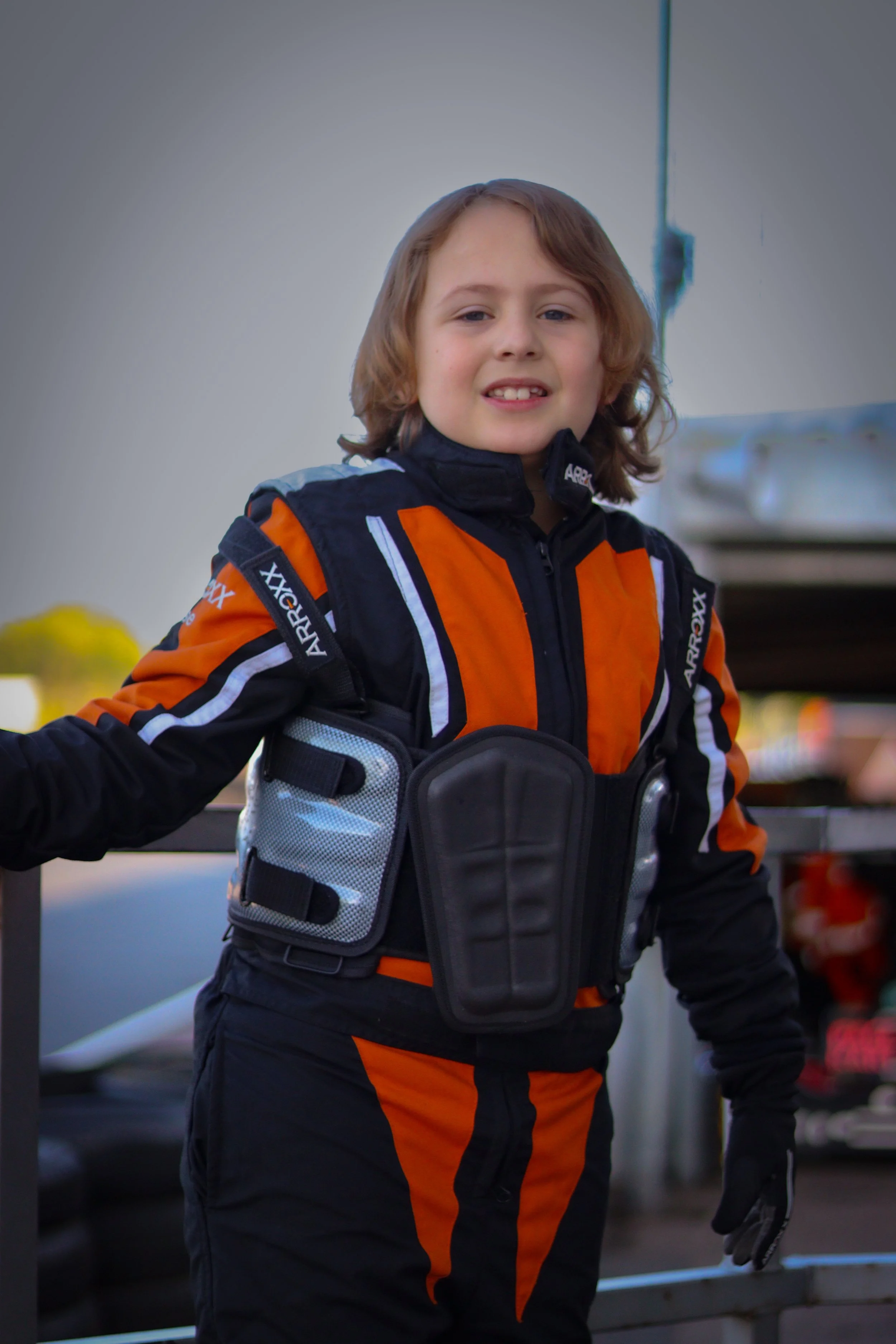 Young boy in race suit and body armor standing outdoors.