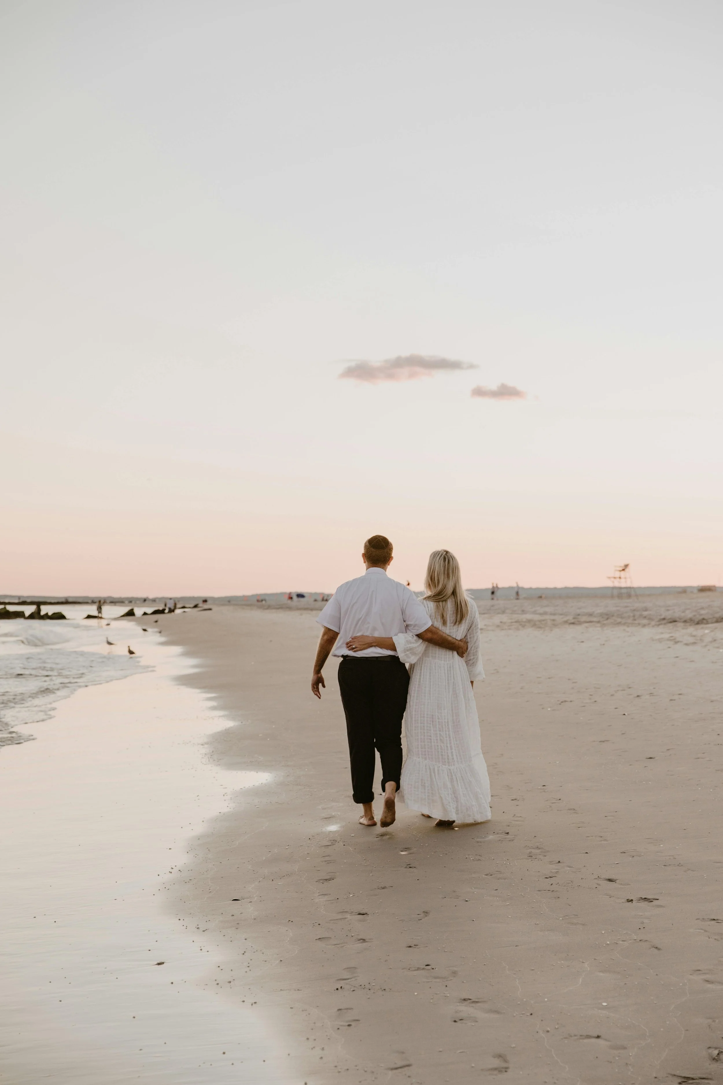 A couple walking hand in hand along the beach at sunset, with gentle waves and a lifeguard tower in the distance.