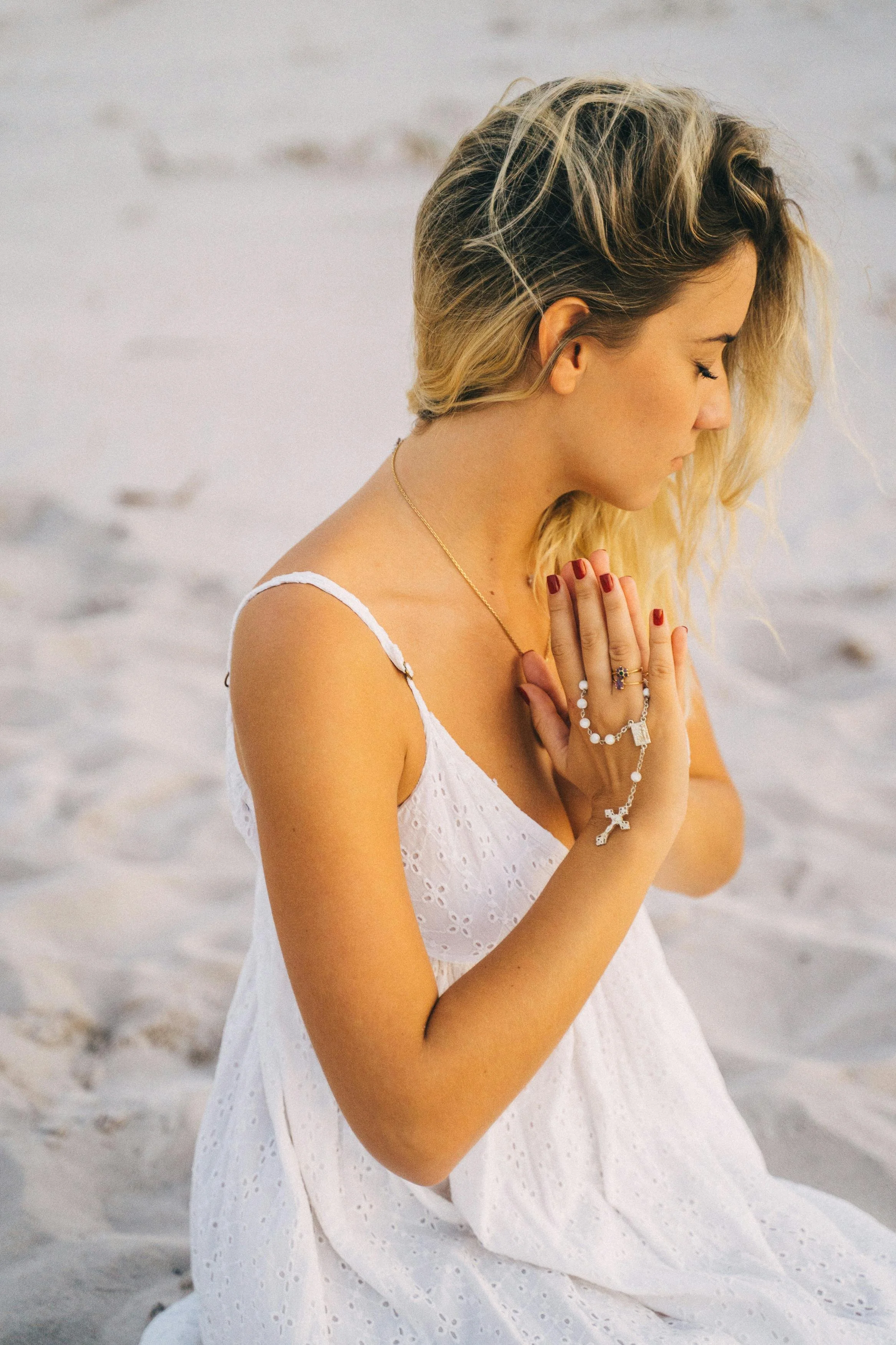 A woman with blonde hair in a white dress, kneeling on the beach with her eyes closed and hands pressed together in prayer.
