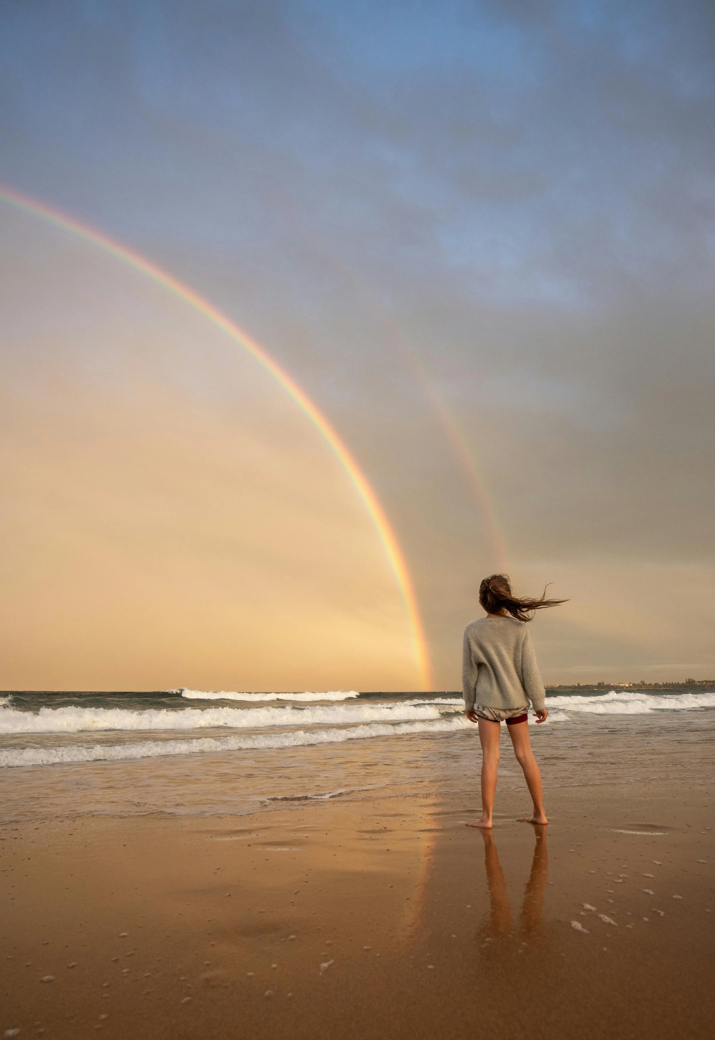 A person standing on a beach at sunset with a colorful rainbow in the sky and waves in the ocean behind them.