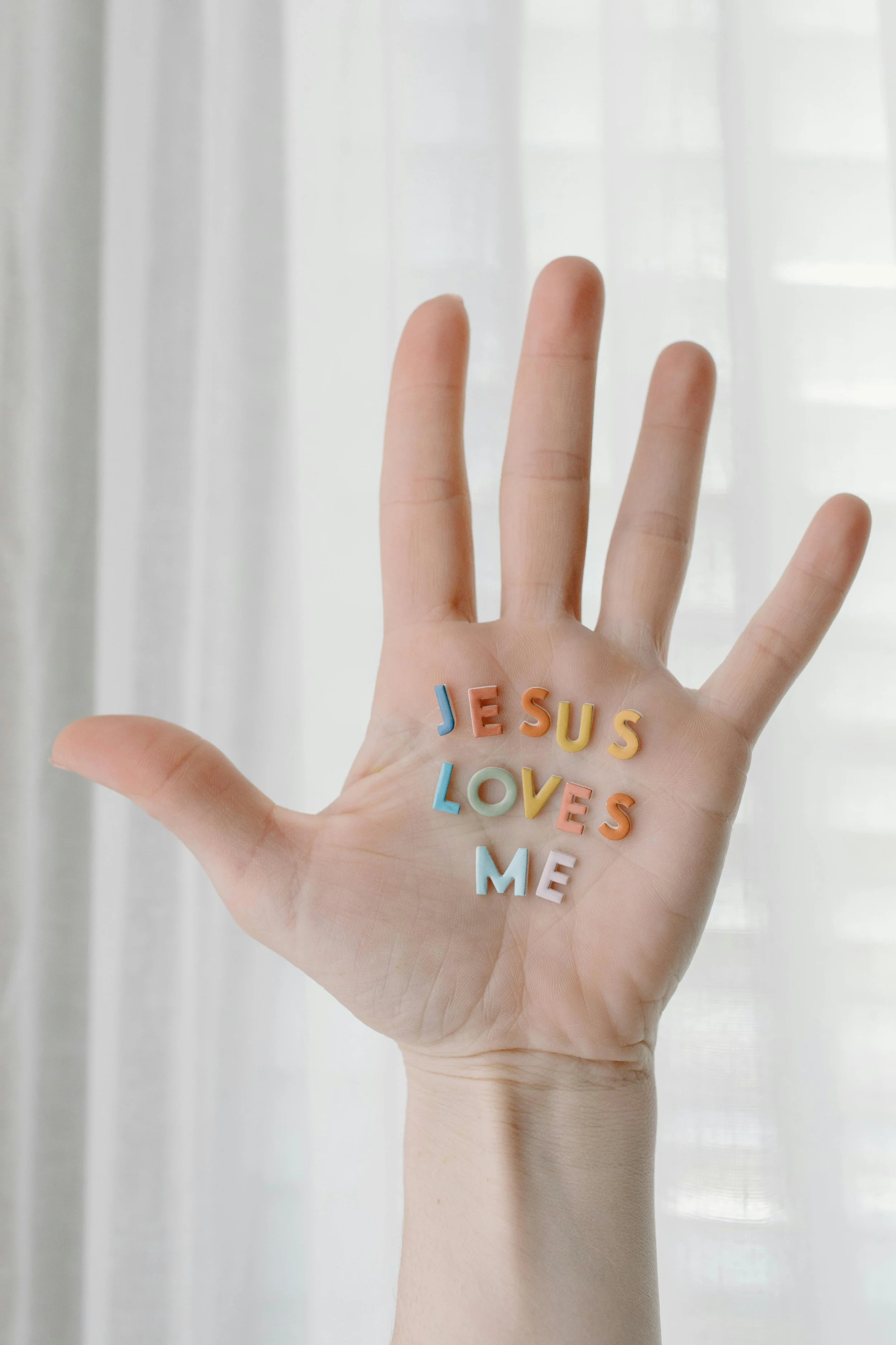 A hand with colorful letter stickers spelling 'JESUS LOVES ME' is displayed in front of a white curtain.