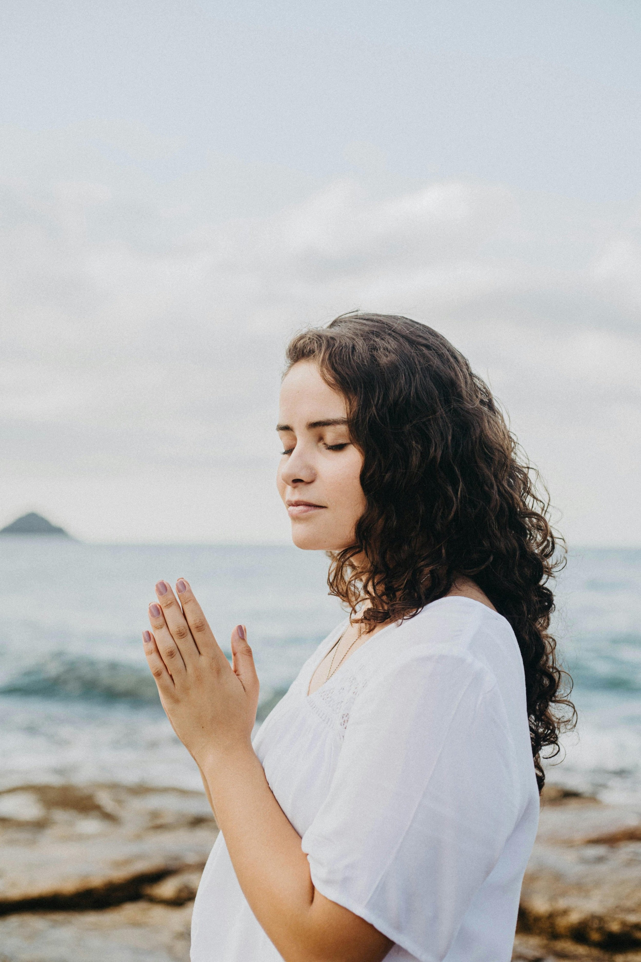 A young woman with curly brown hair and closed eyes, standing on a beach with her hands in a prayer position near her face, wearing a white shirt, during daytime with an overcast sky.