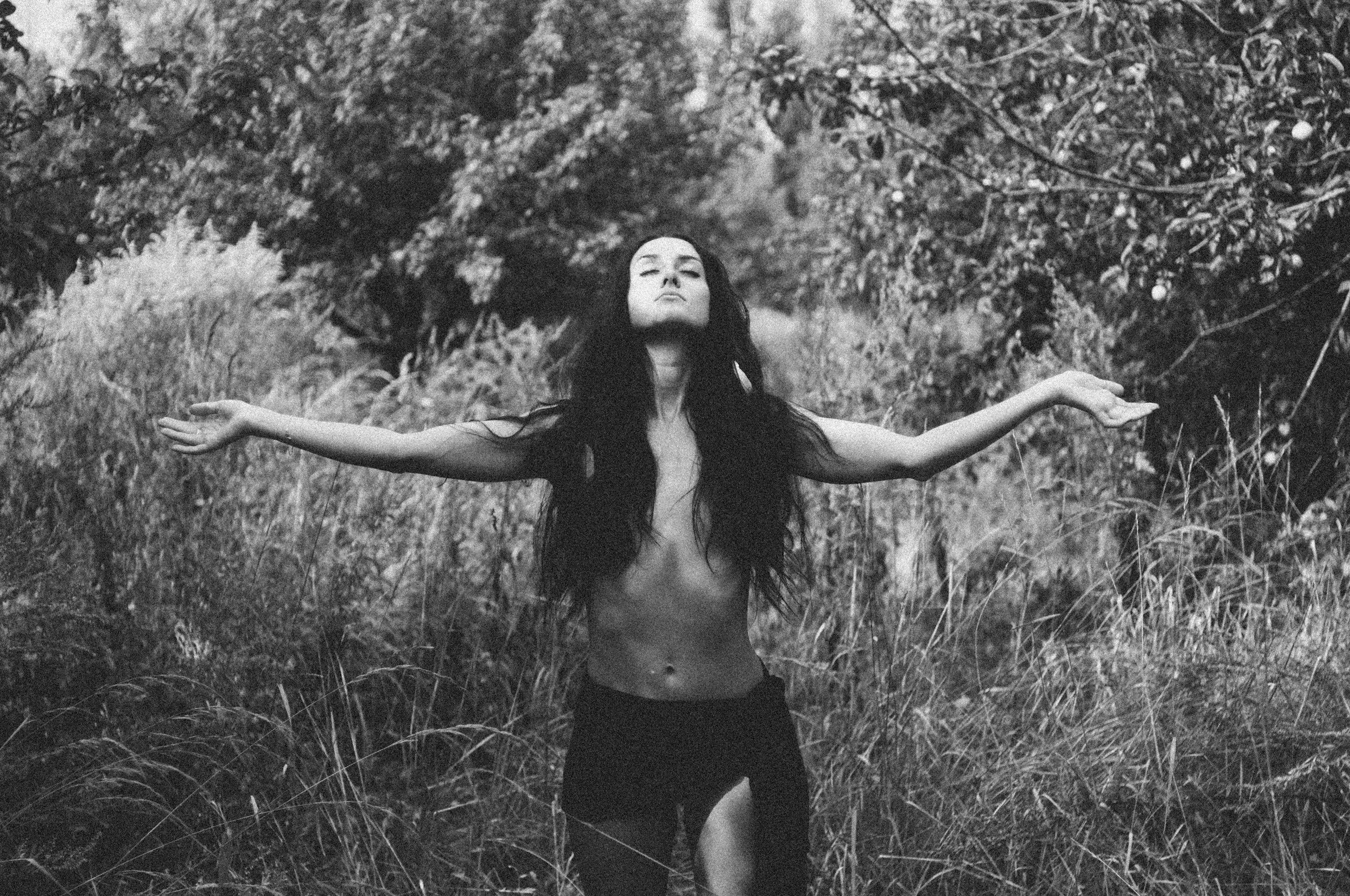 A woman with long dark hair standing outdoors in a natural setting with her arms outstretched and eyes closed.