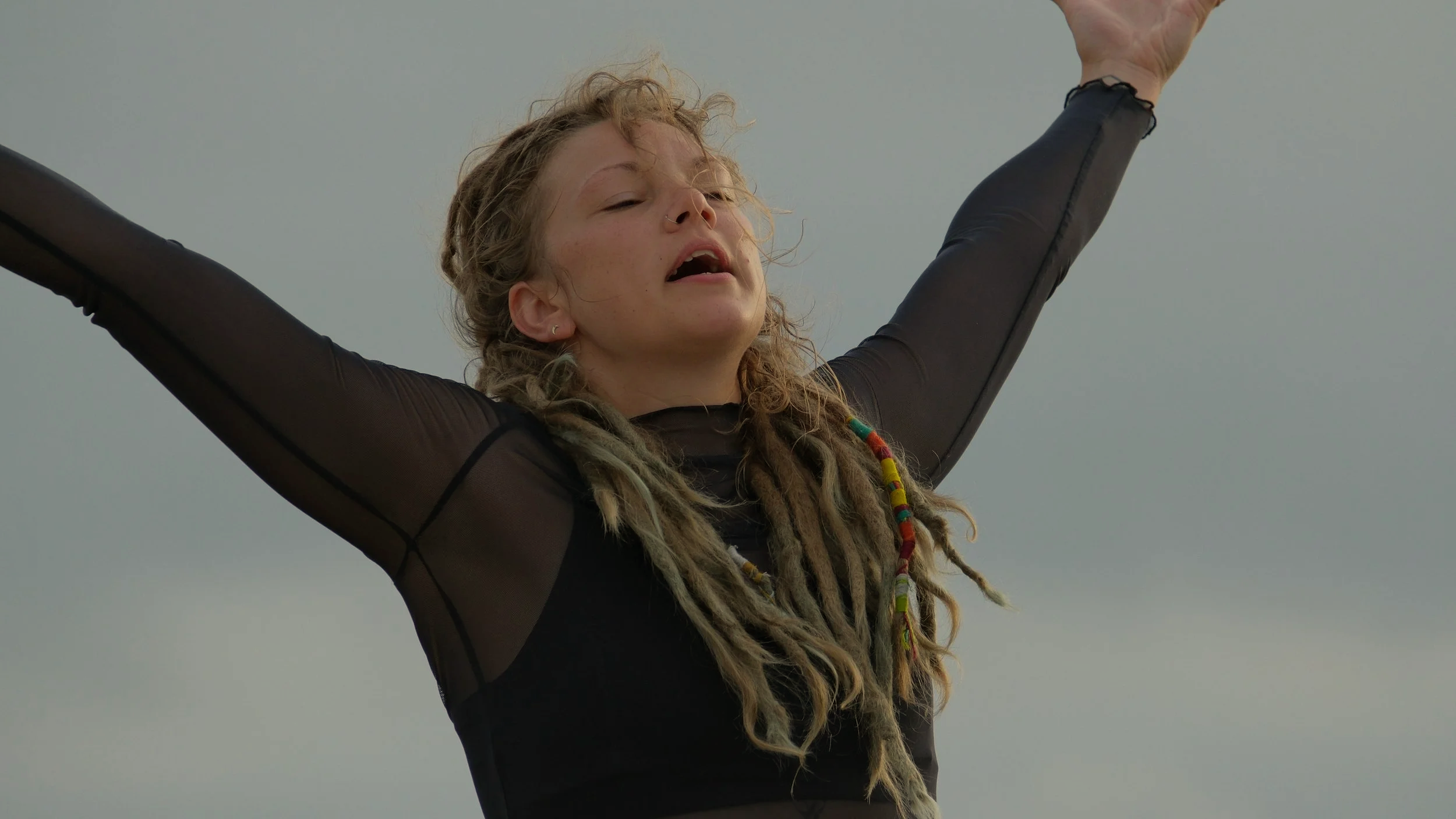 A young woman with light skin and dreadlocks wearing a black sheer long-sleeve top with her arms raised and eyes closed, standing outdoors under a cloudy sky.