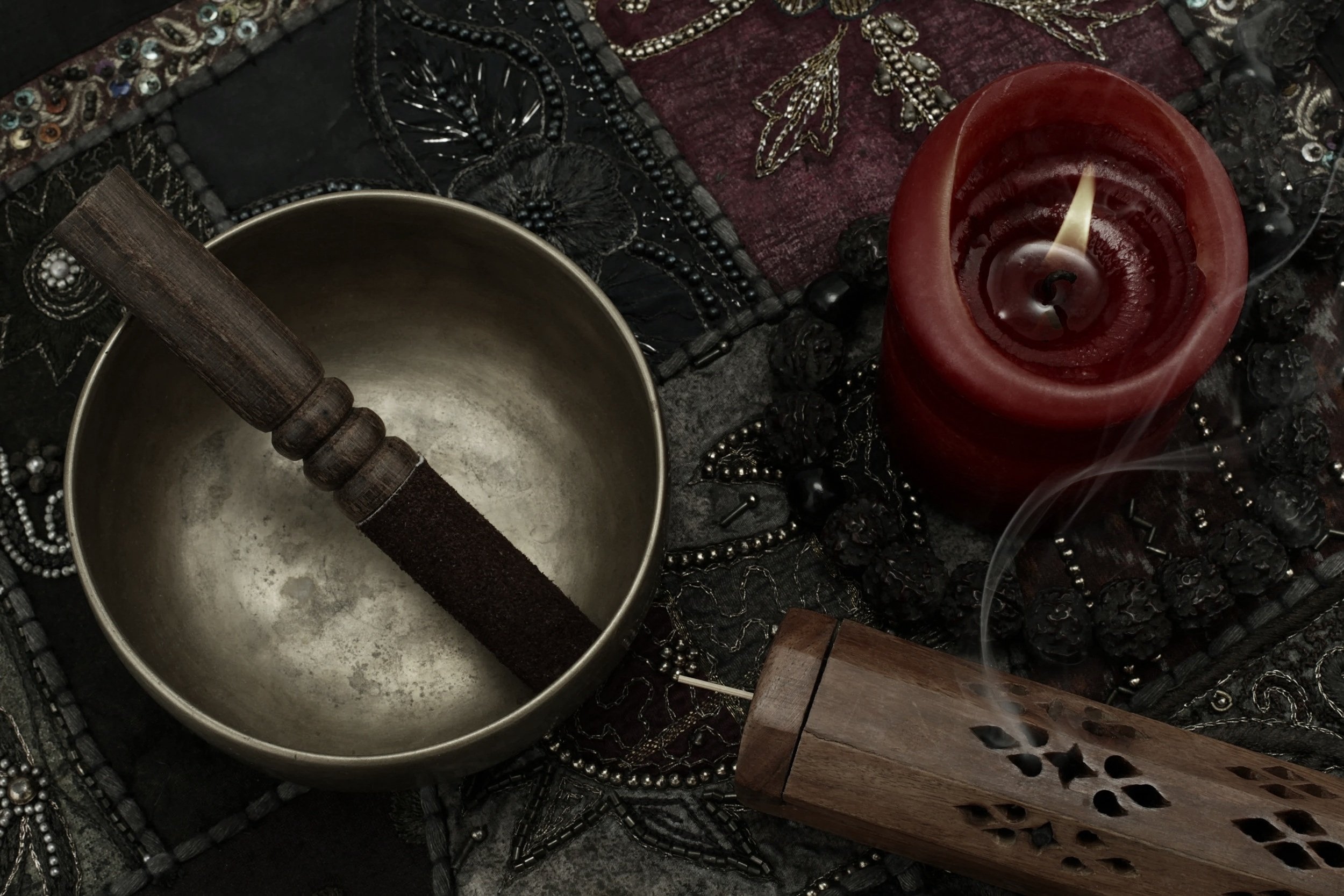A collection of musical and decorative objects including a singing bowl with a wooden striker, a lit red candle, and various jewelry and beaded accessories on a dark embroidered fabric surface.
