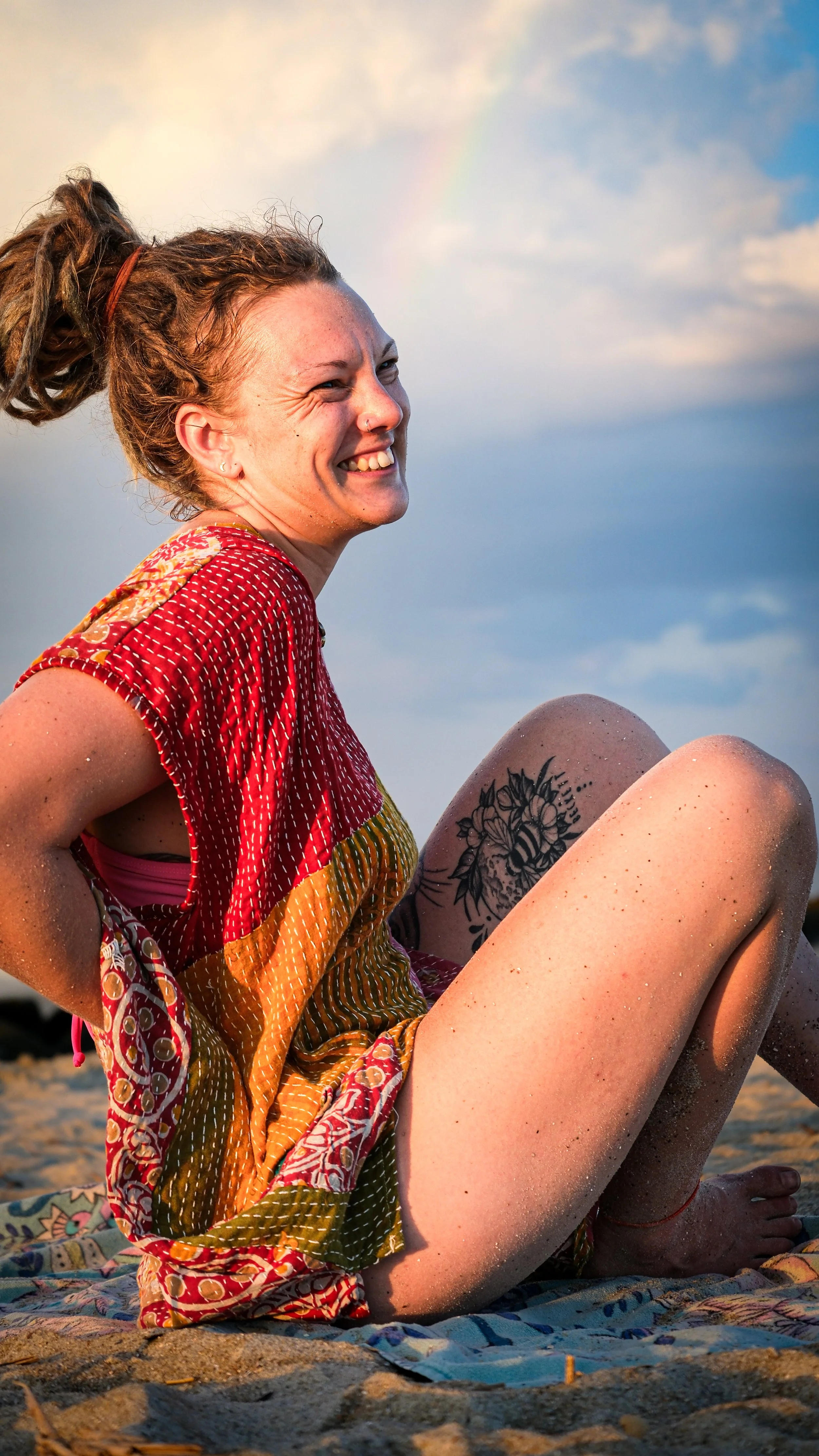 A woman with dreadlocks smiling while sitting on a sandy beach during sunset, with a tattoo of flowers on her thigh, and a rainbow faintly visible in the sky.
