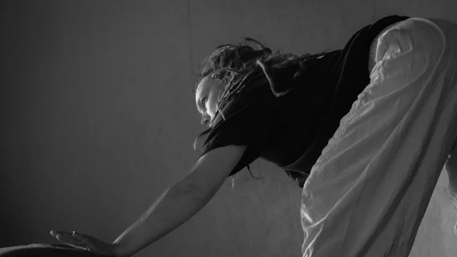 A woman in a black shirt and white skirt leaning forward, resting her hands on the floor, in a room with plain walls.