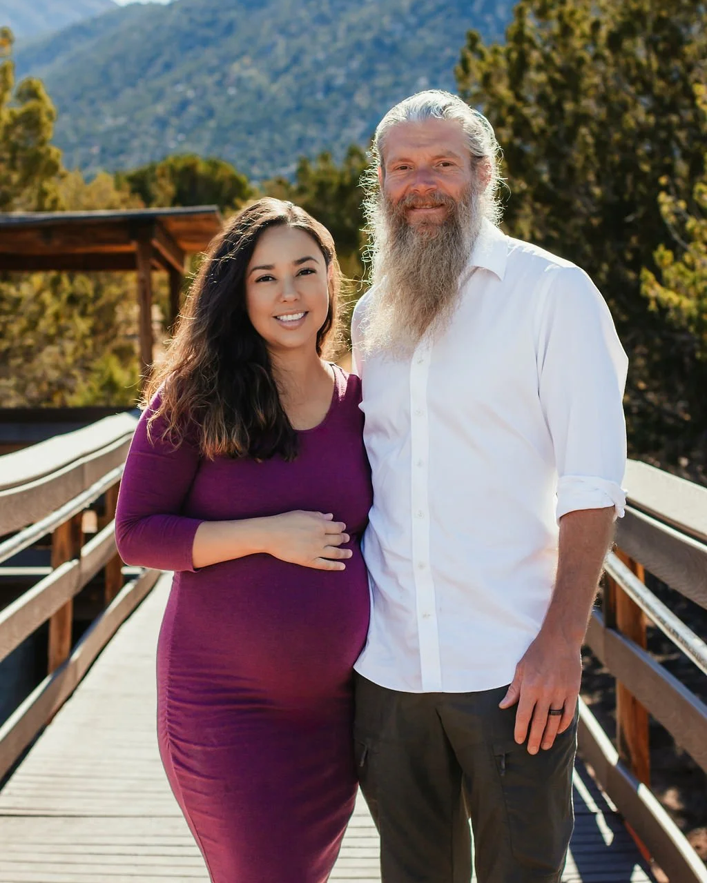 A smiling young woman with long dark hair and a white man with a long gray beard standing on a wooden bridge outdoors with green trees and mountains in the background.
