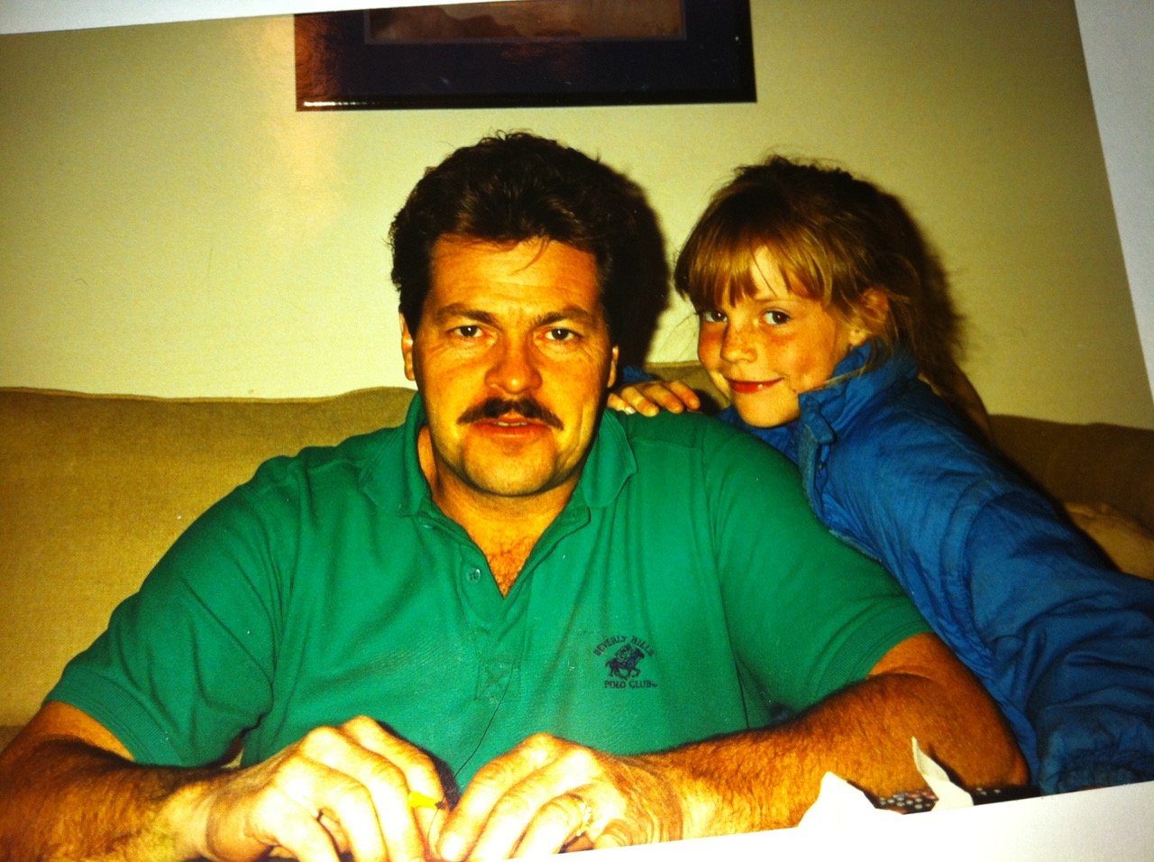 A man with dark hair and a mustache, wearing a green polo shirt, sitting on a couch with a young girl with light brown hair and a blue jacket, smiling and resting her arm on his shoulder.