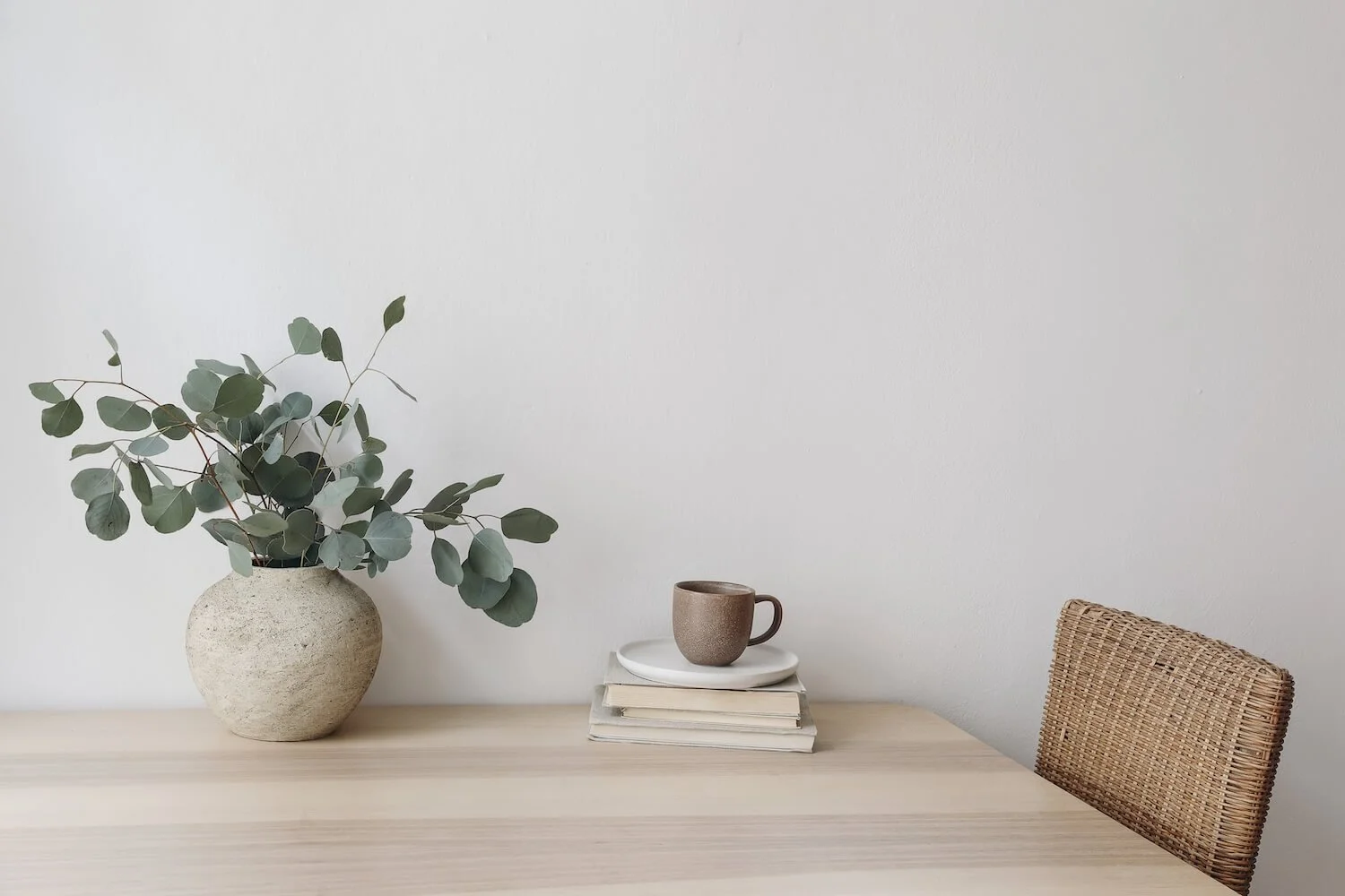 A beige tables with a vases with eucalyptus leaves, a coffee cup, and a stack of books, against a light-colored wall, with a woven chair on the right.