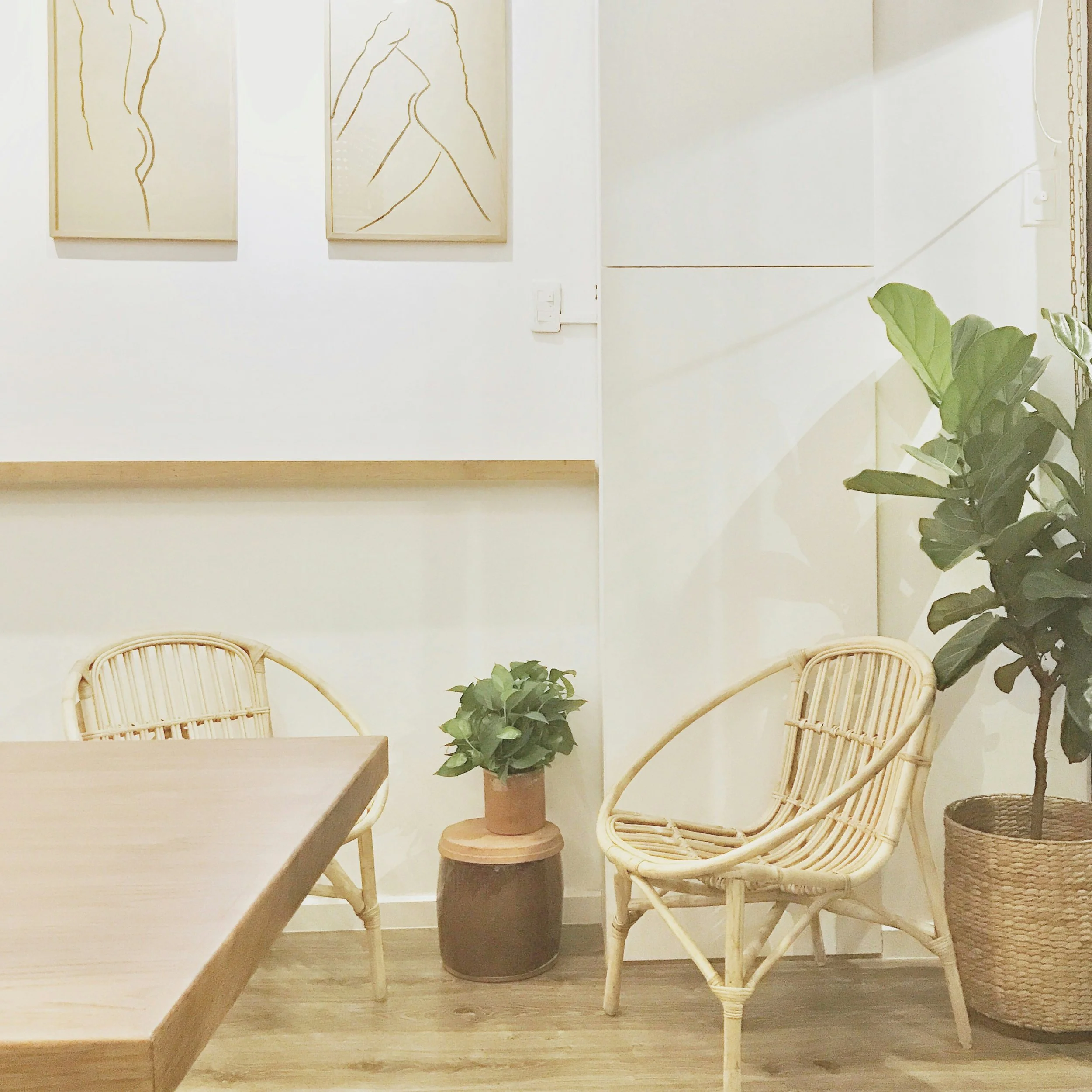 Interior of a room with two light-colored rattan chairs, a small round side table with a potted plant on it, and three minimalist line art portraits on the white wall.