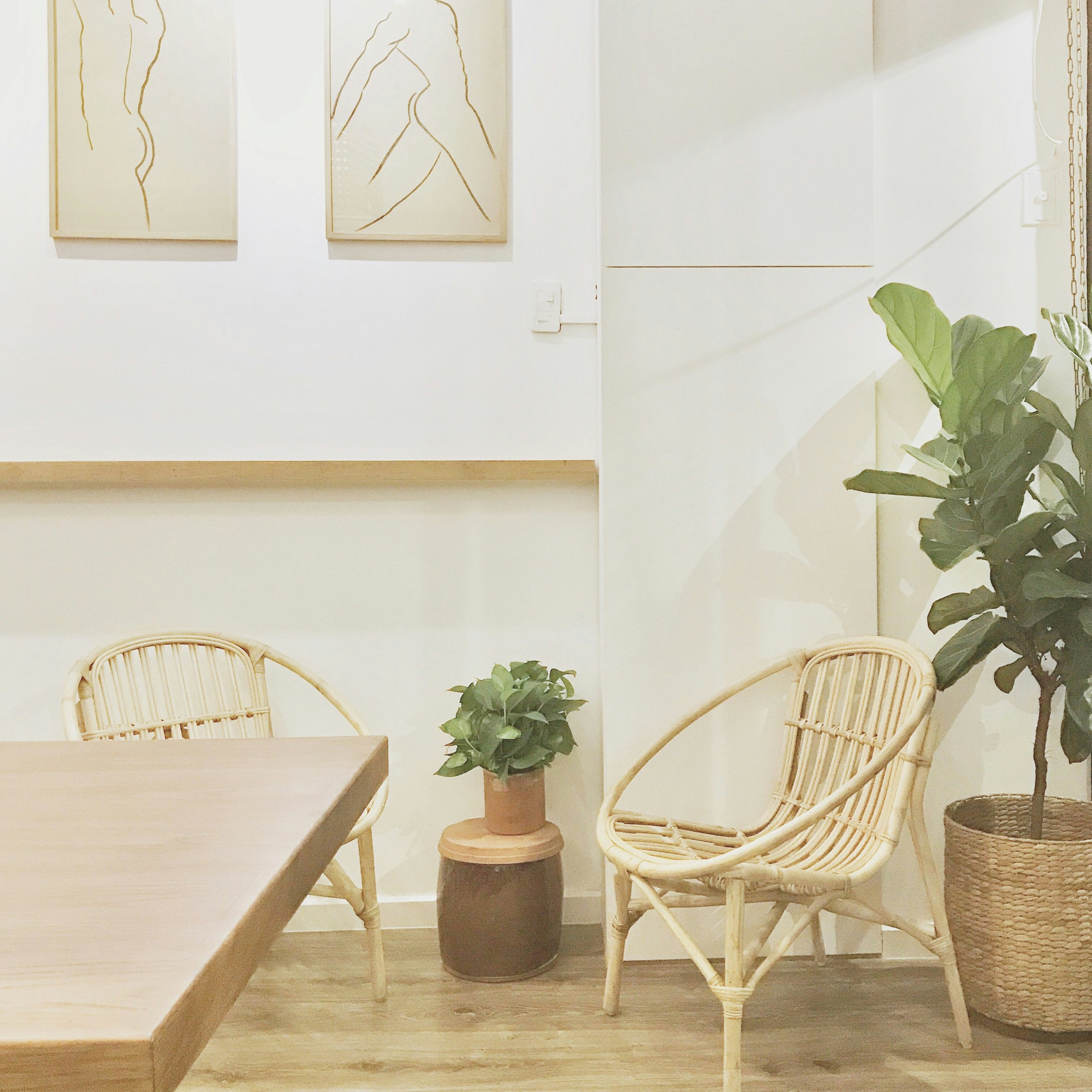 A cozy corner with two wicker chairs, a small wooden table, and a potted plant, all against a white wall decorated with minimalist line art of human figures.