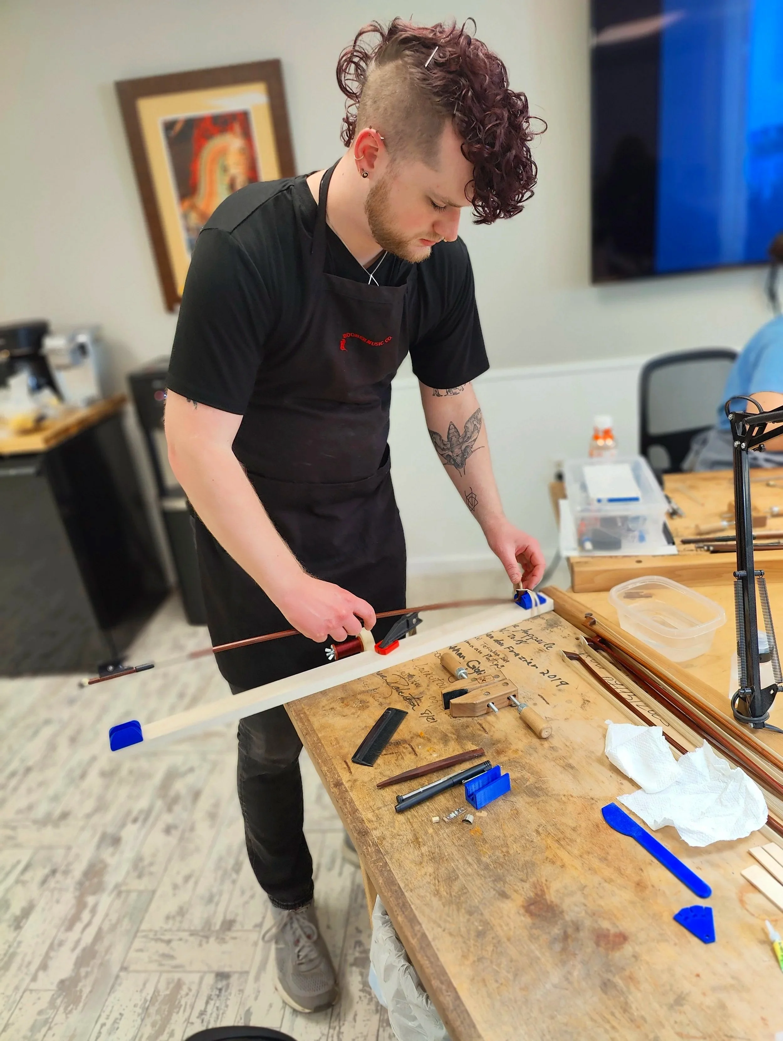 A person with curly purple hair and tattoos rehairing a violin bow at a wooden table, with various tools and materials around.