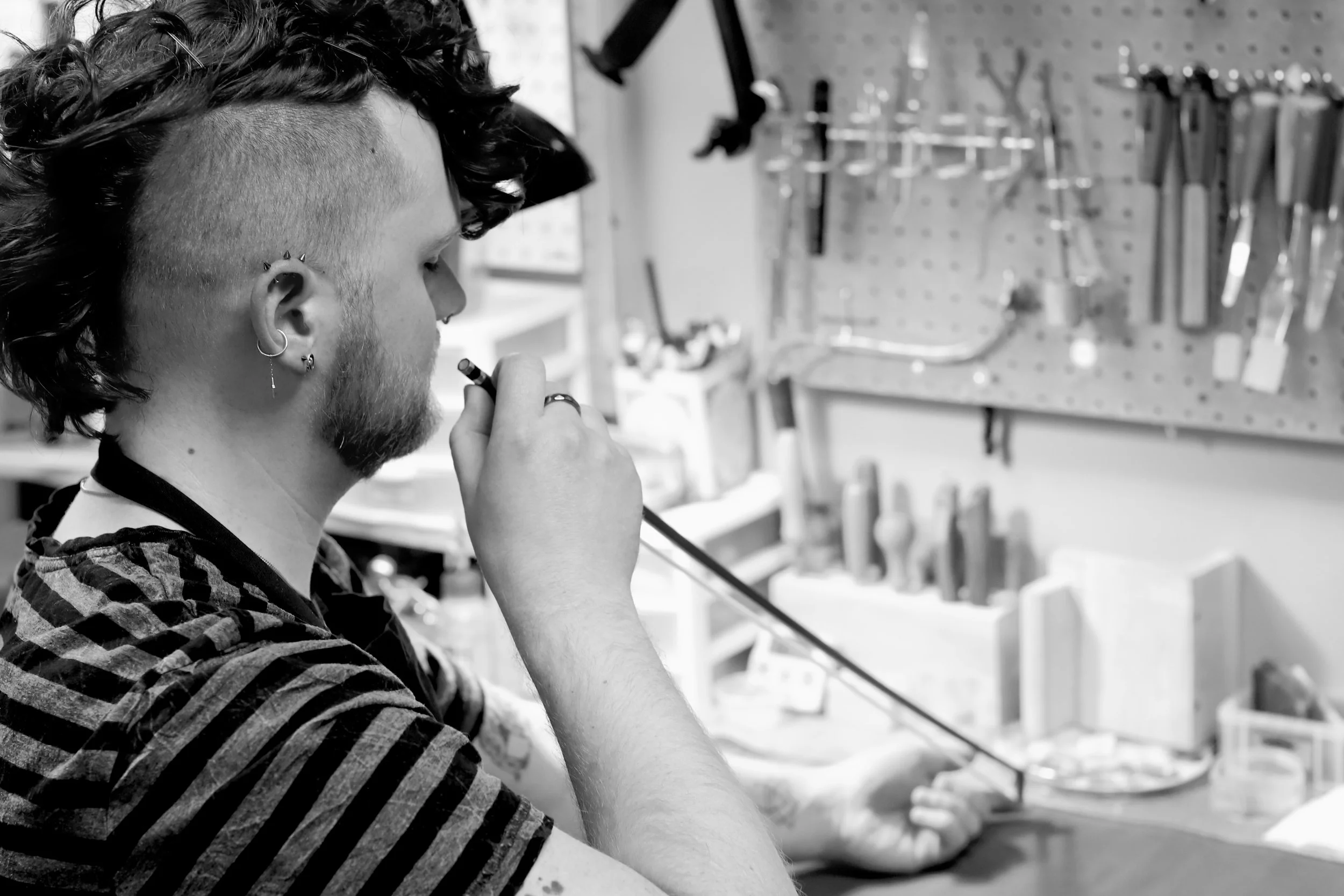 A person with a curly, mohawked hairstyle and multiple earrings, wearing a striped shirt, is assessing a violin bow for warping in a workshop with organized tools on a pegboard in the background.