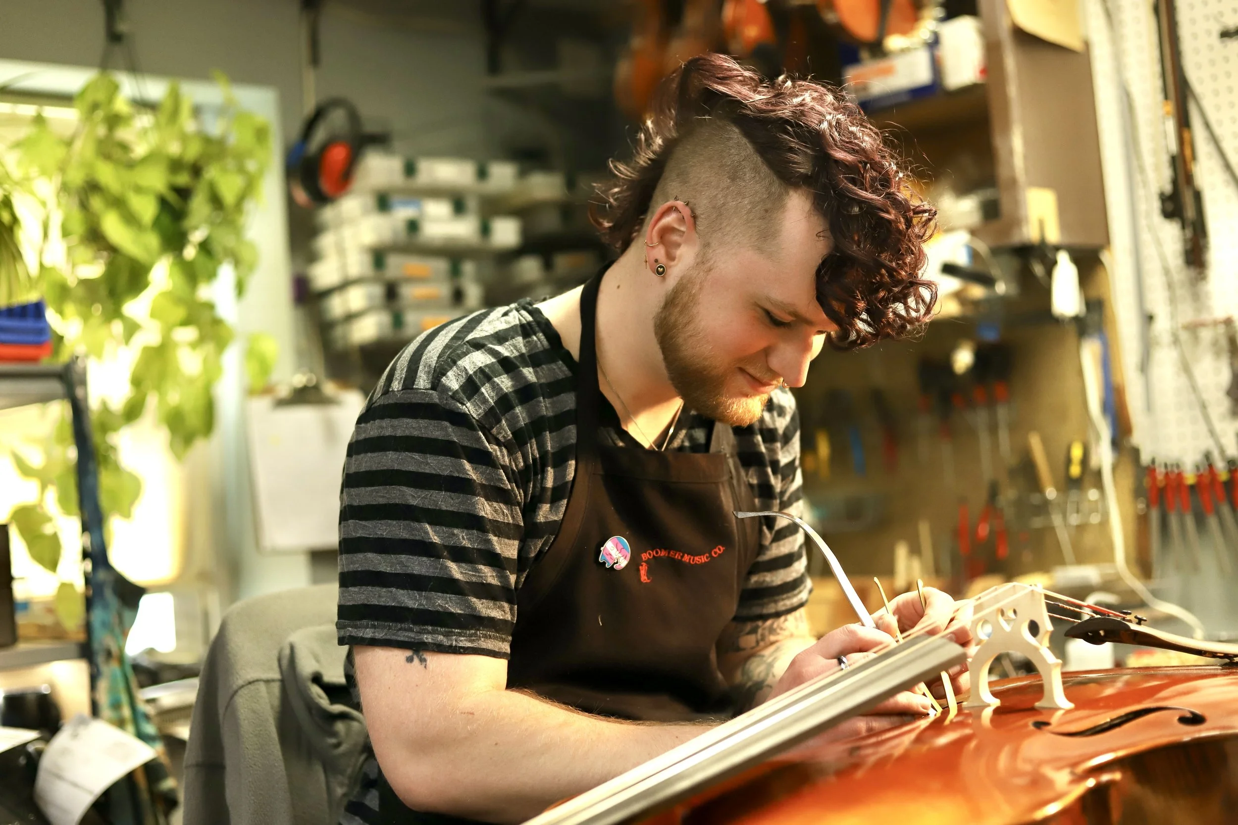 A person in their 30s with a curly mohawk and tattoos working on a cello in a workshop, wearing a black apron with a Boomer Music logo, surrounded by tools and equipment.