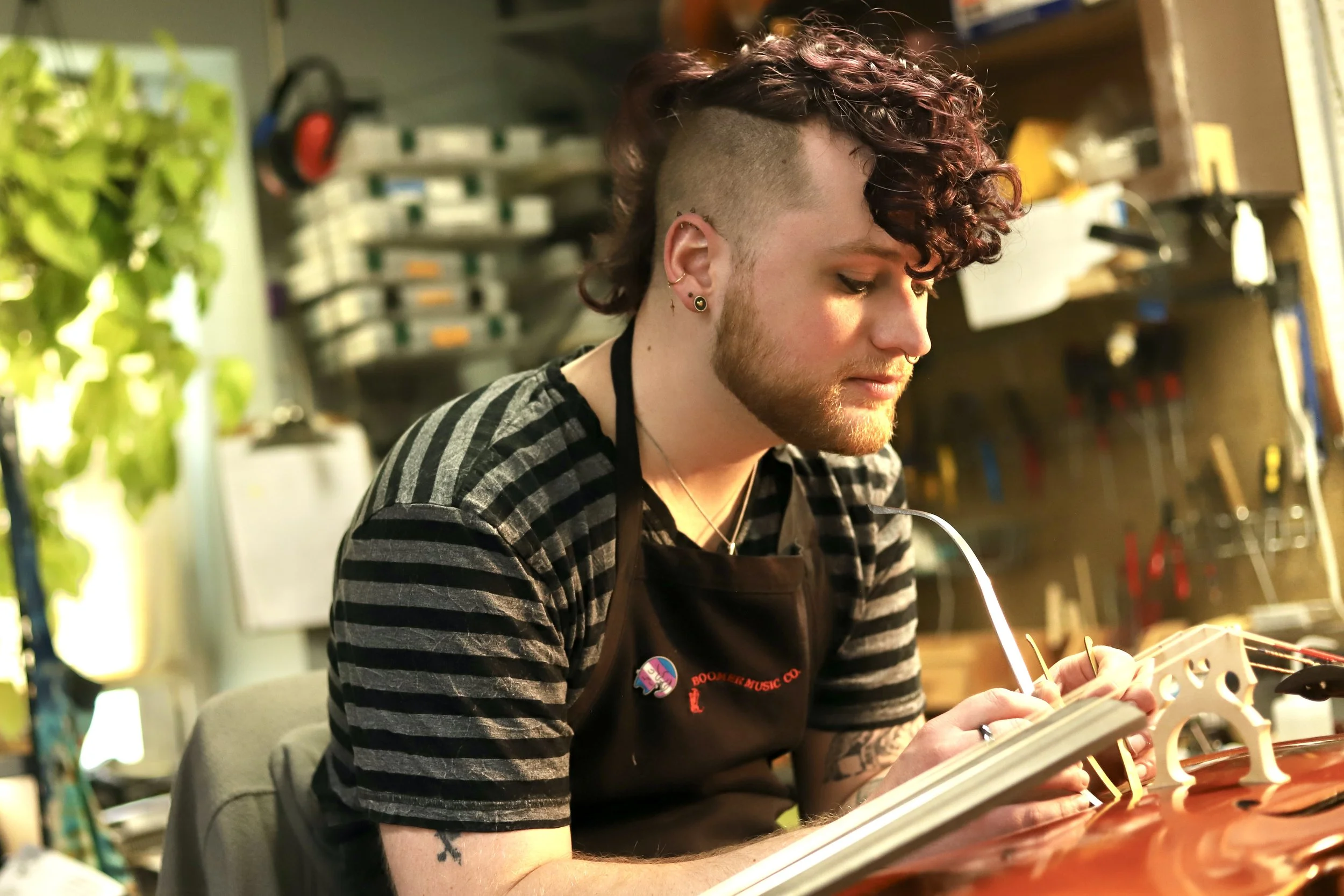 A person in their 30s with a curly mohawk and tattoos working on a cello in a workshop, wearing a black apron with a Boomer Music logo, surrounded by tools and equipment.