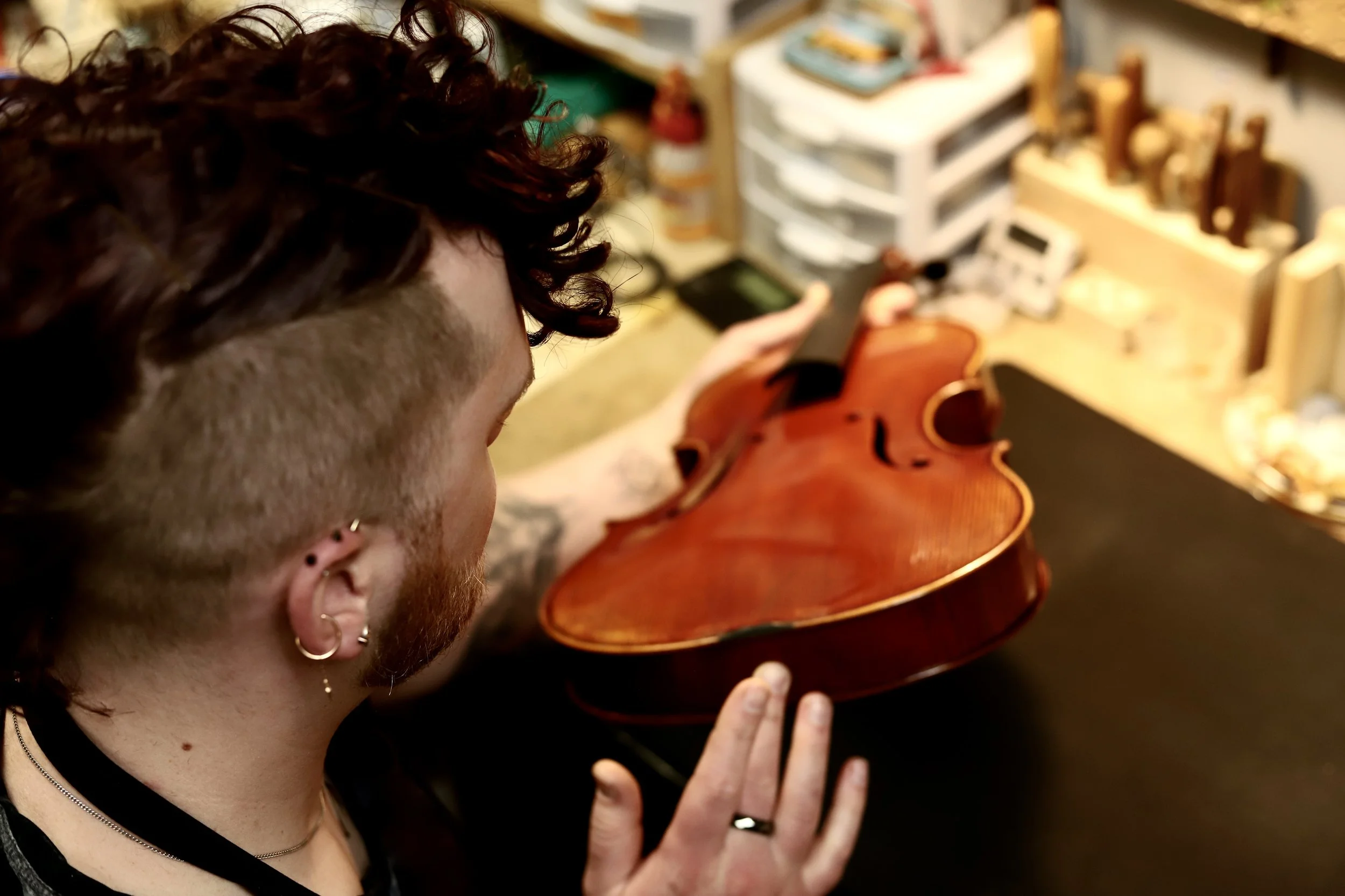 Person with curly hair inspecting a viola in a workshop with shelves and various carpentry tools.