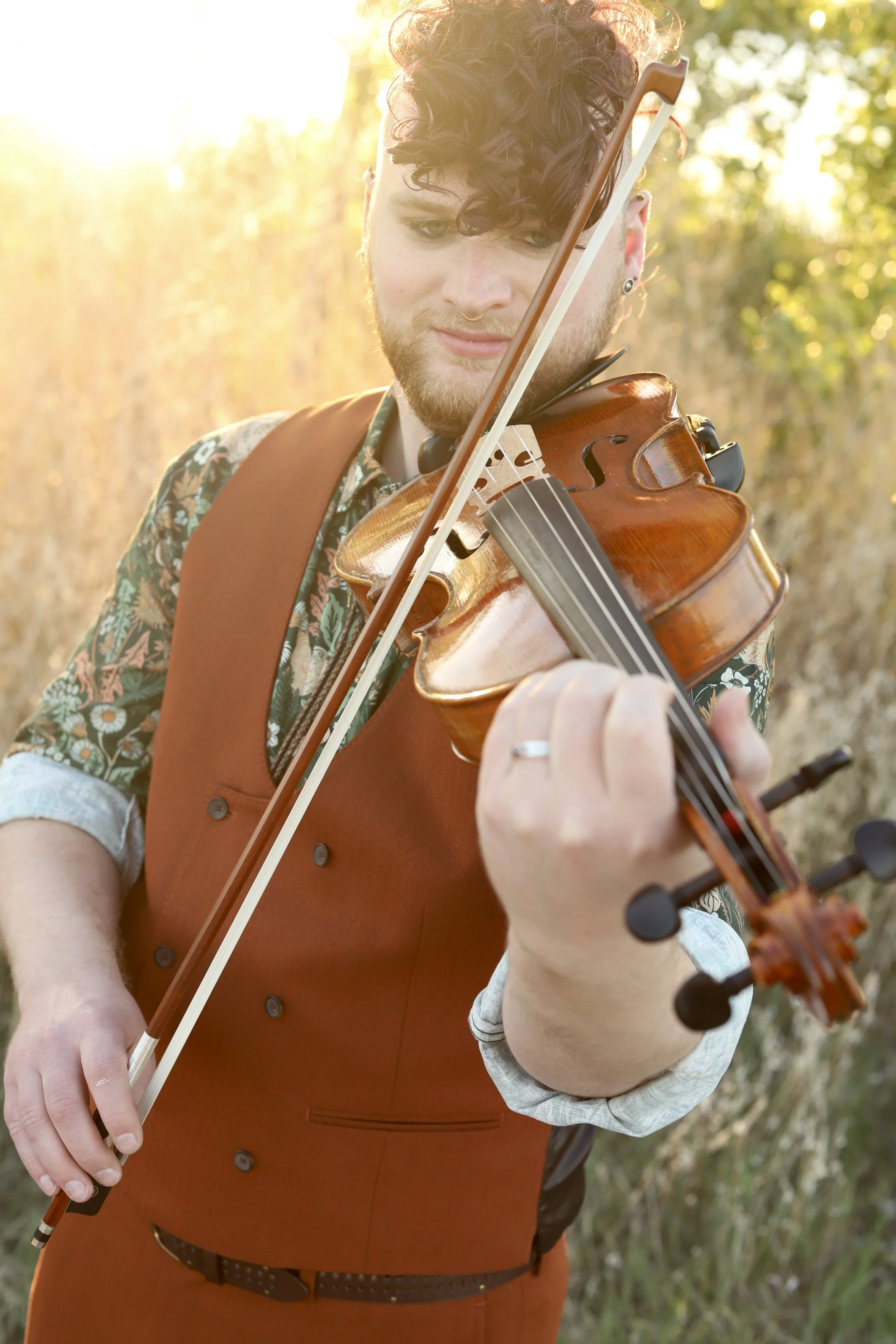 A person playing the violin outdoors during sunset, with sunlight glowing in the background and trees around.