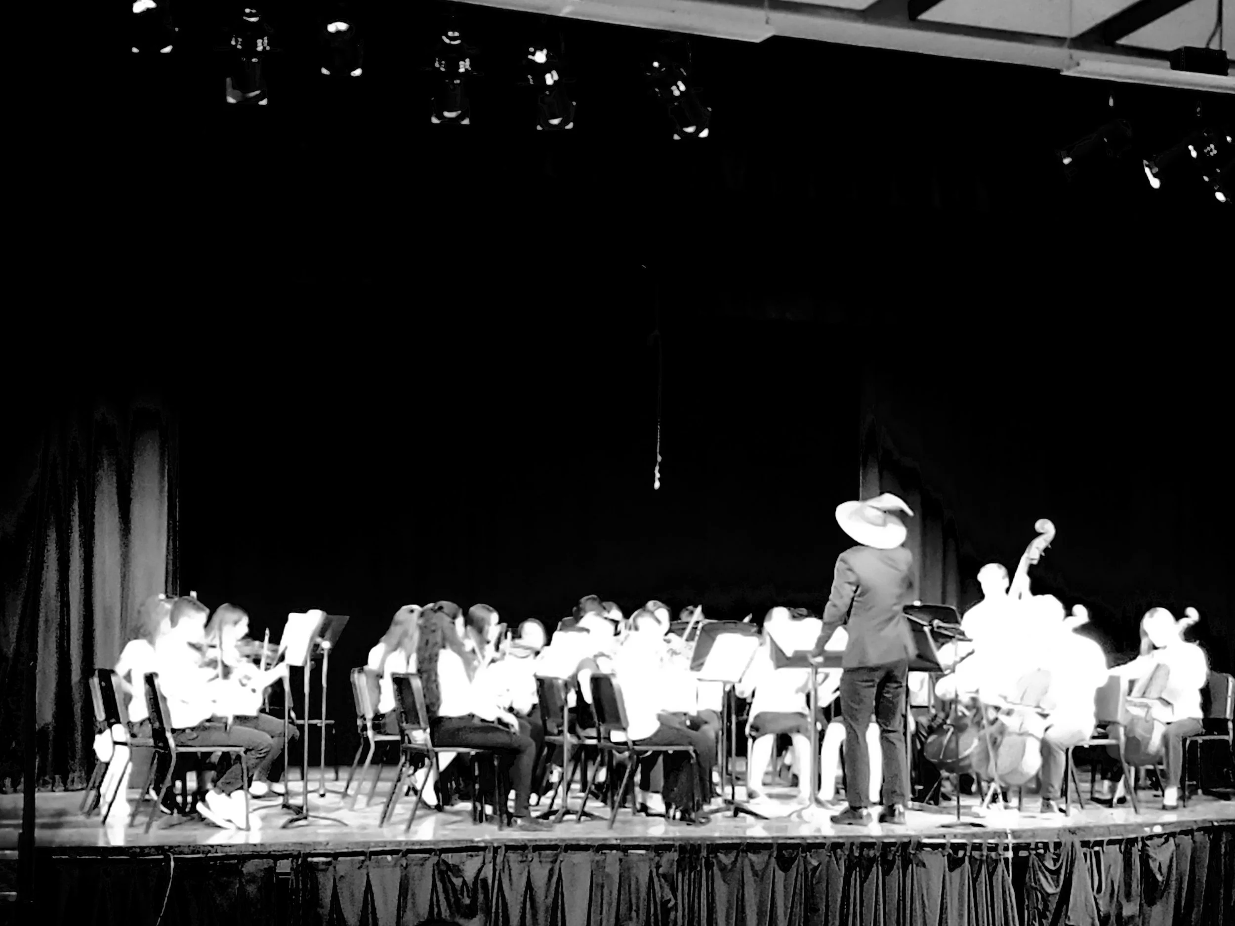 A large group of children on stage, seated in rows, playing musical instruments, directed by a conductor wearing a Gandolf-style wizard hat, in a school auditorium with curtains and stage lighting.