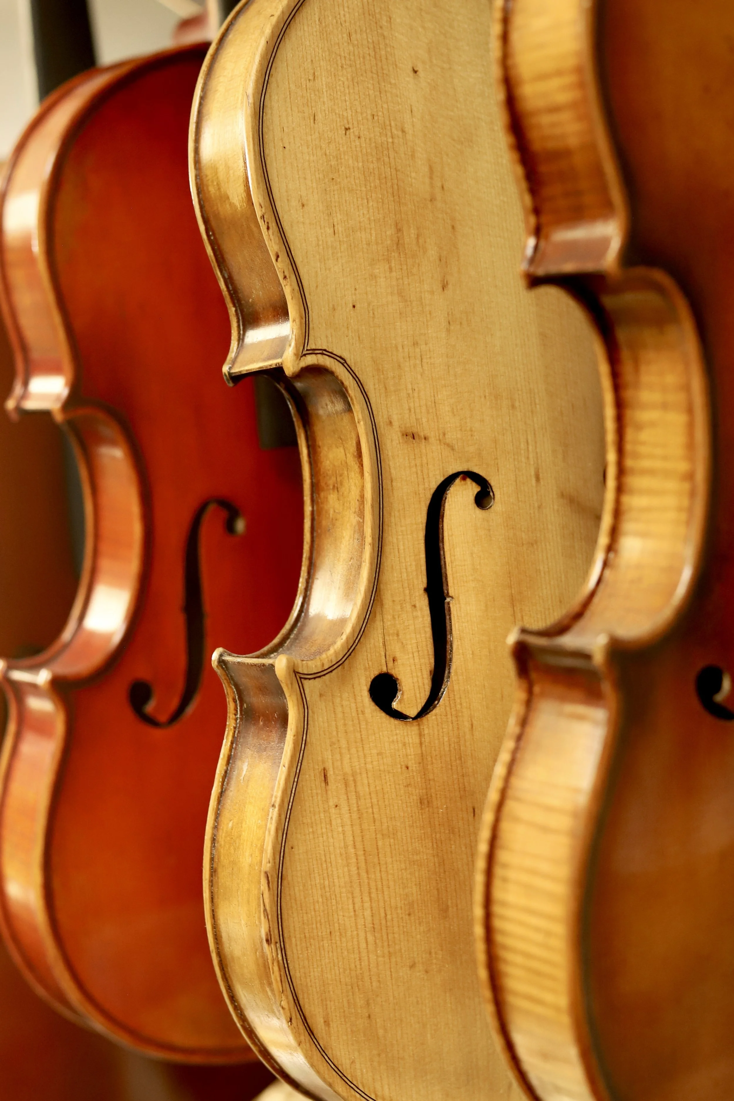 Close-up view of three violins, focusing on their curved wooden bodies and f-holes, showing natural wood finish and craftsmanship.