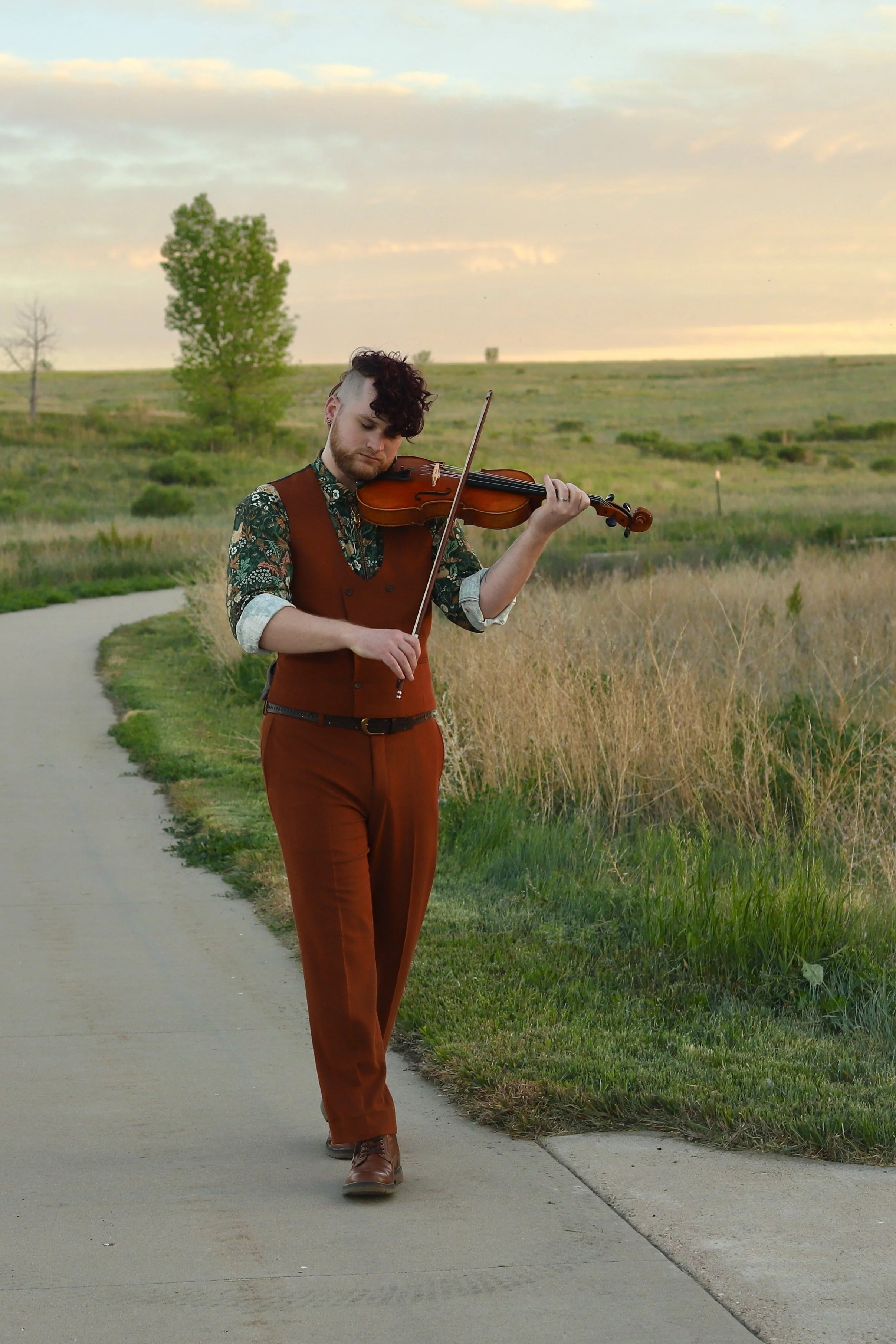 A person with a curly mohawk, dressed in a floral shirt, brown vest, and brown pants, playing the viola outdoors on a sidewalk in a grassy field during sunset.