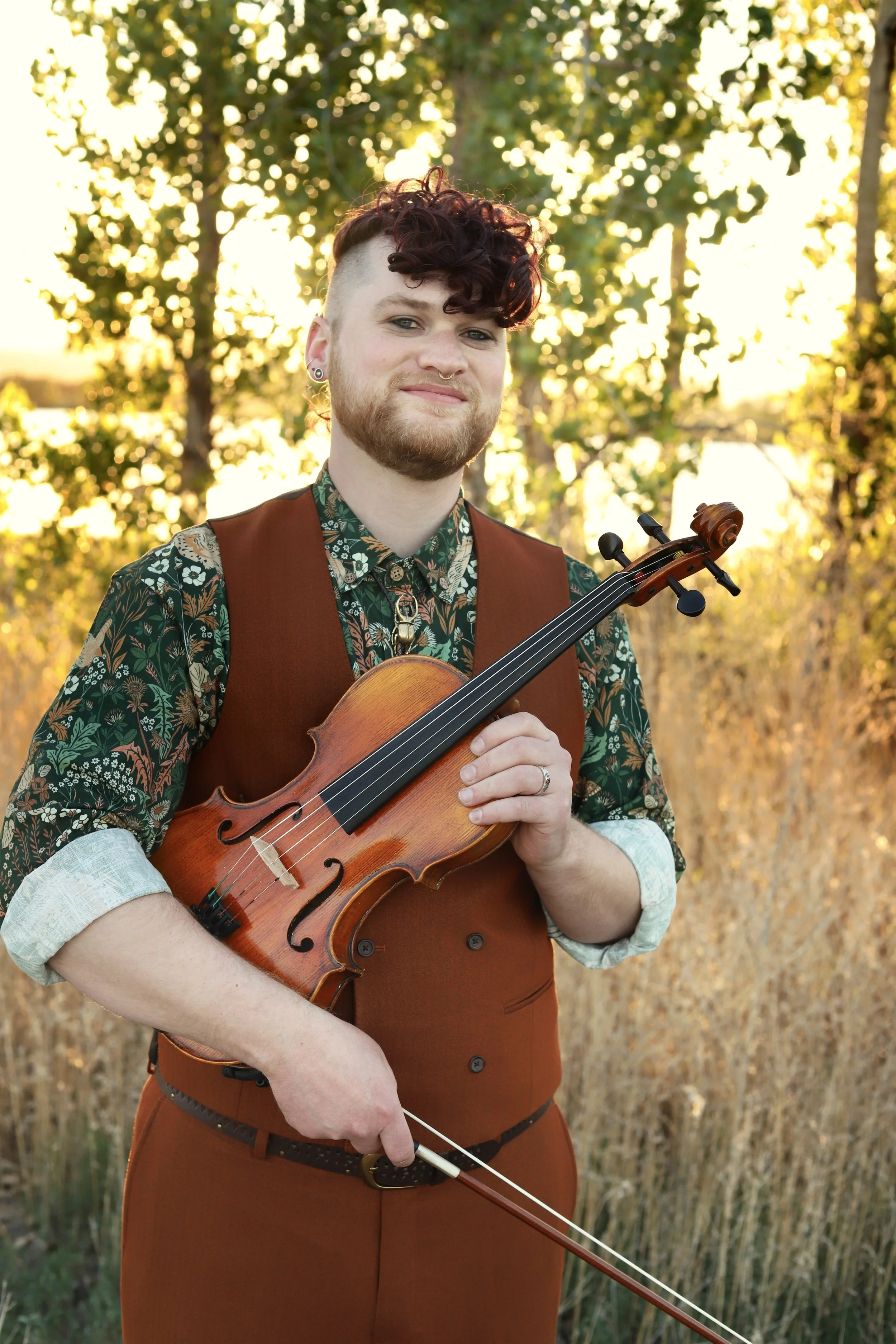 A person with curly purple hair and a beard playing a viola outdoors during golden hour, surrounded by trees and tall grass.