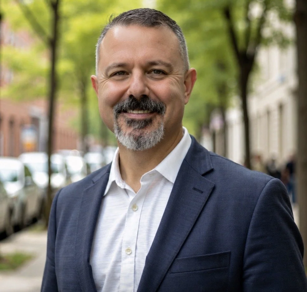 A well-groomed, middle-aged man with short dark hair, a beard with gray, wearing a navy blazer and white shirt, smiling outdoors on a city street with trees, cars, and buildings in the background.