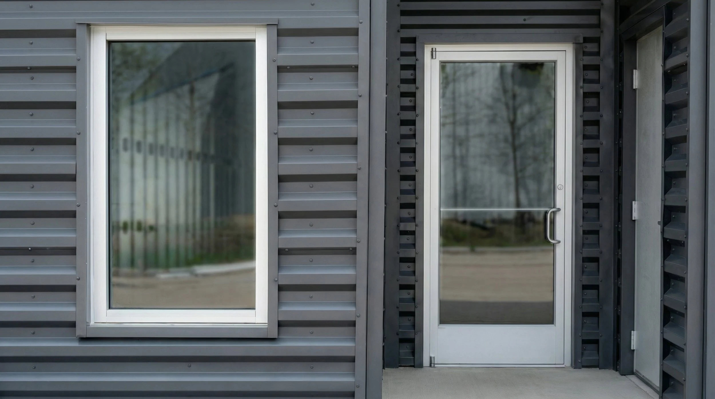 A building exterior with gray metal siding, large glass windows, and a glass door with a metal handle, reflecting some trees and sky.
