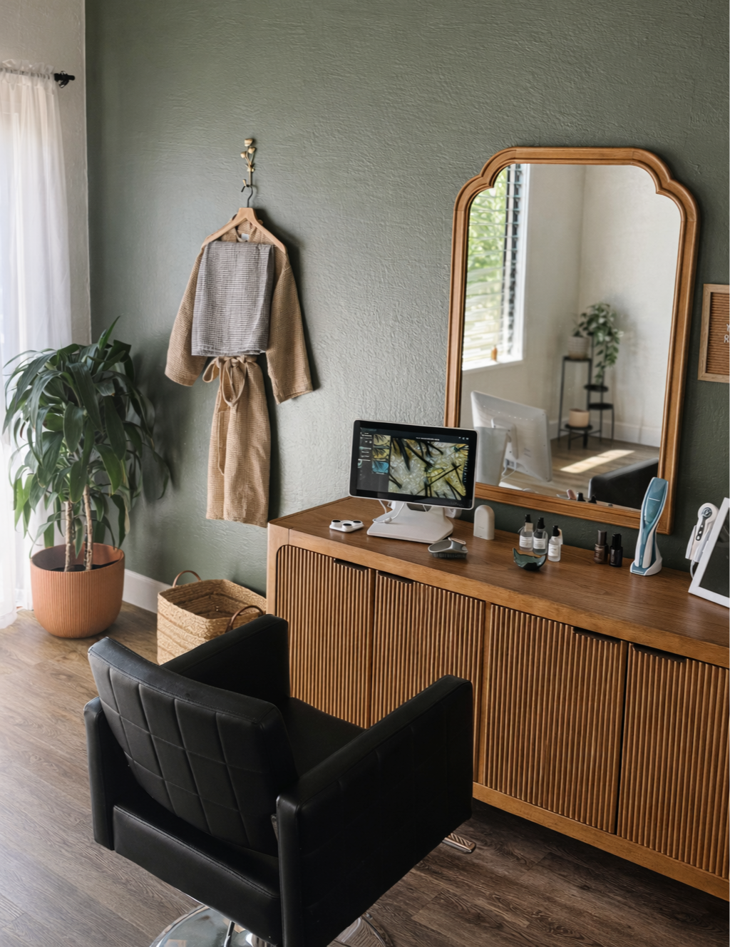A modern vanity area with a large mirror, a computer monitor, skincare products, and an electric toothbrush. A black chair in front, a green wall, a large potted plant, and a bathrobe hanging on a hook.
